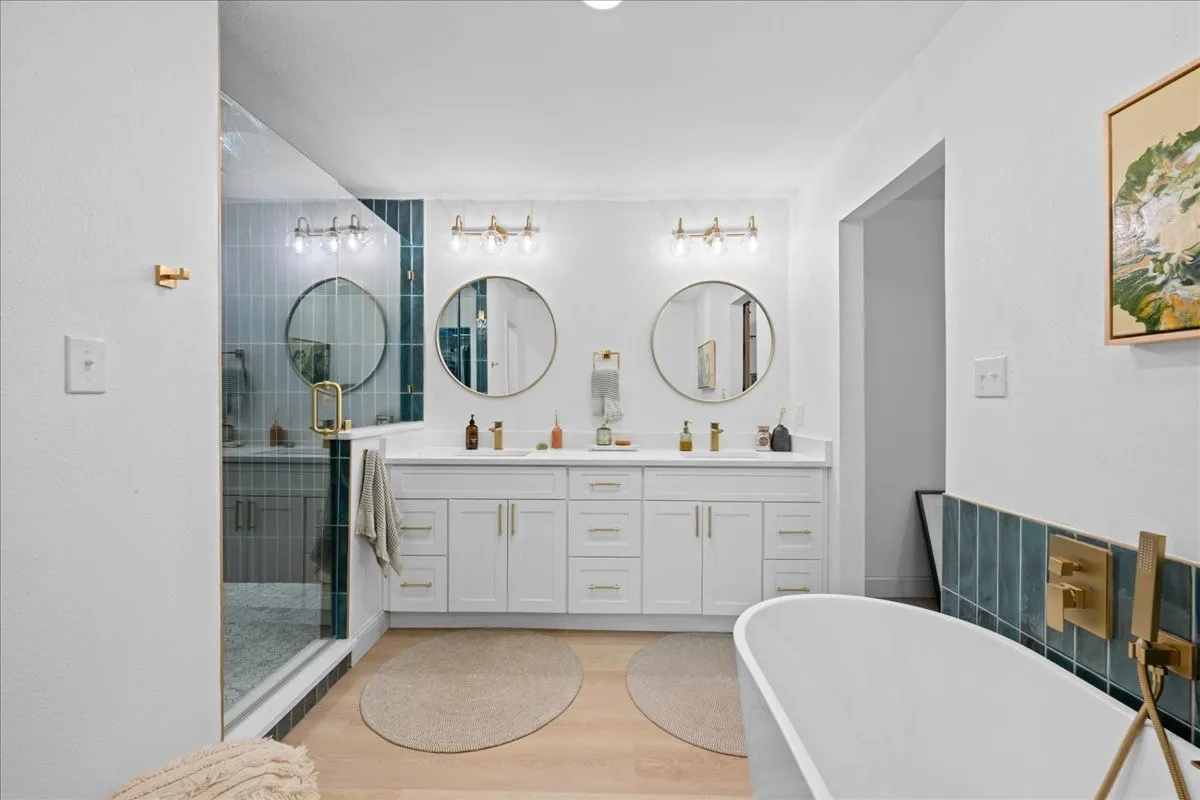 Dual vanity with white cabinetry and circular mirrors, glass-enclosed shower with tile surround, and a freestanding soaking tub