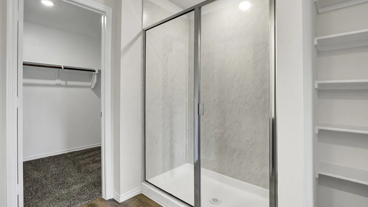 Walk-in shower with clear glass enclosure, built-in shelving, wood-finish flooring, and a carpeted closet with hanging rod and shelving
