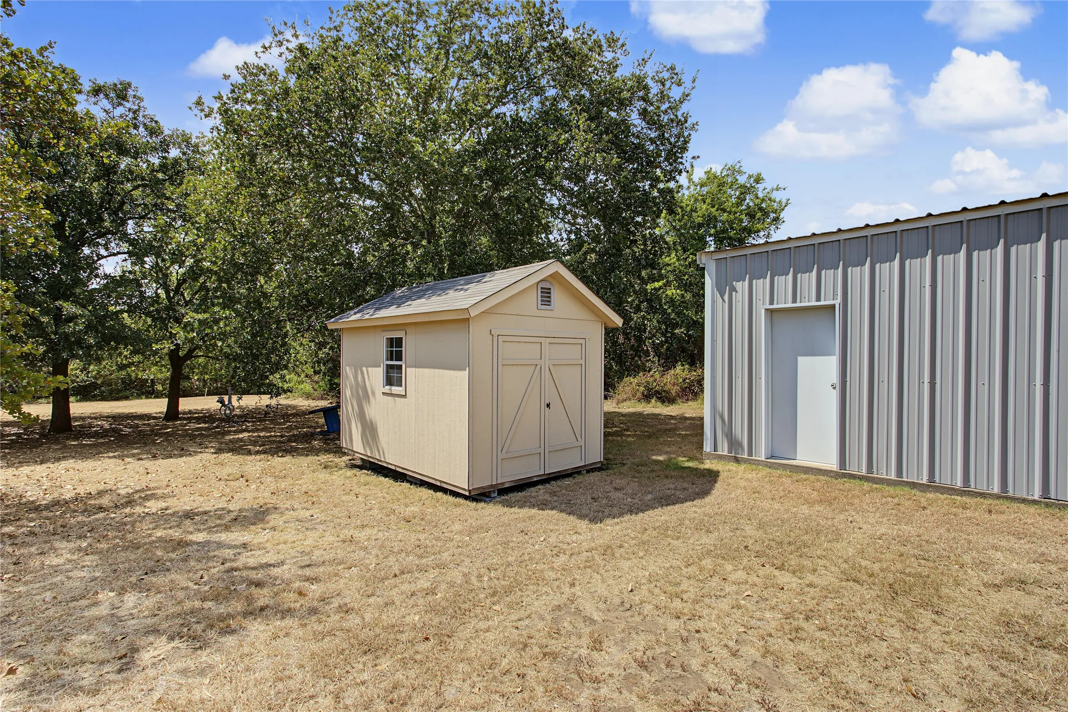 Detached storage shed with a single window and gabled roof, alongside a large corrugated metal outbuilding featuring a single entry door
