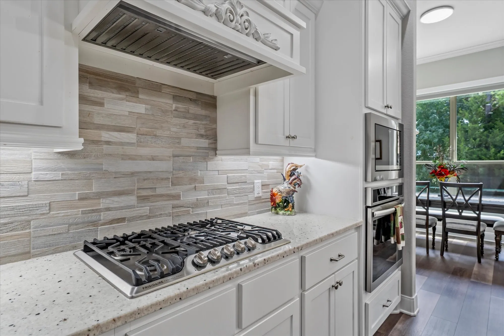 White cabinetry, integrated gas cooktop, light-toned stacked stone backsplash, white speckled countertop, and stainless steel built-in appliances