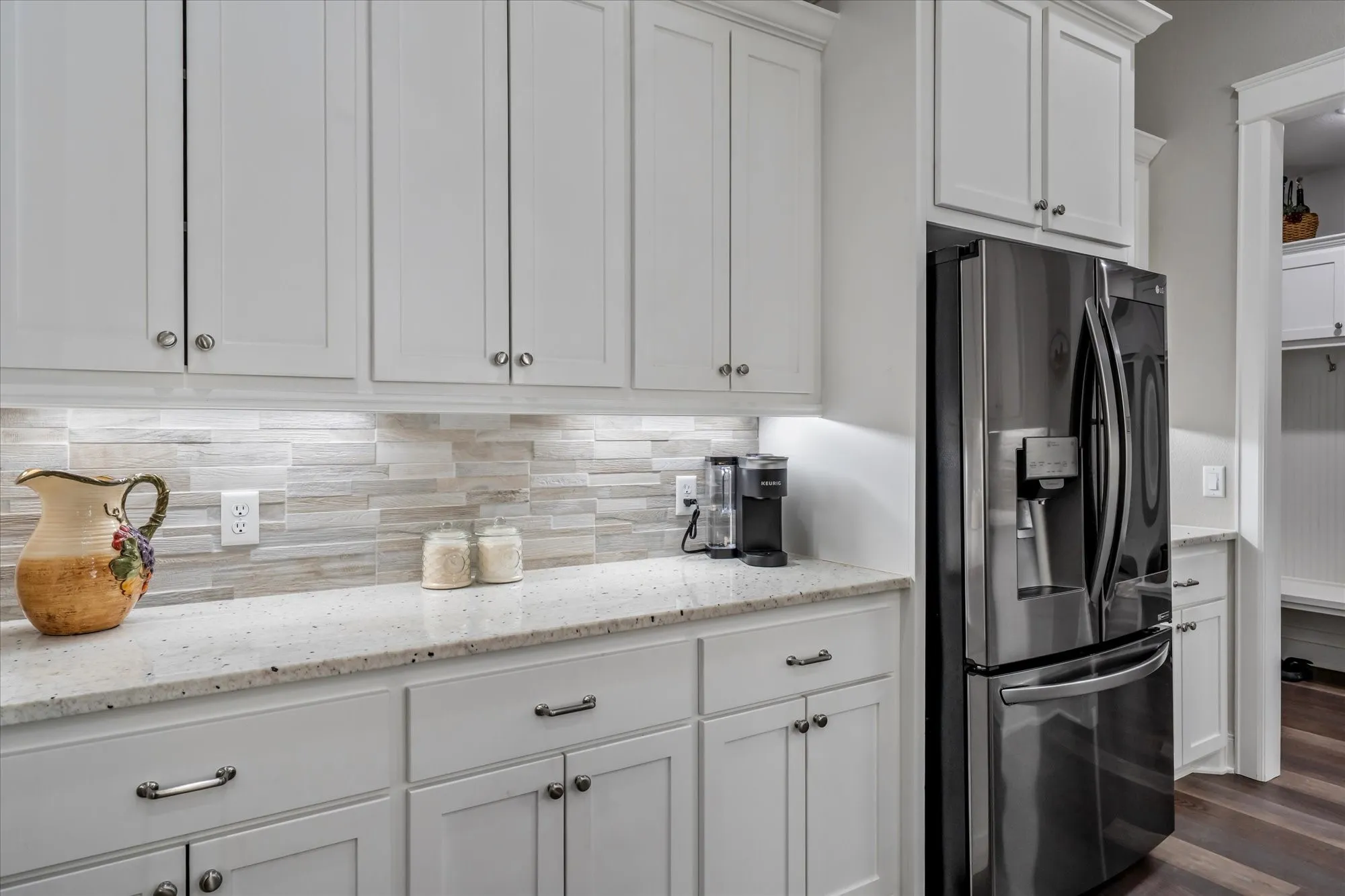 White shaker-style cabinetry, light-toned stone-finish countertops, stainless steel French door refrigerator, and stacked linear tile backsplash