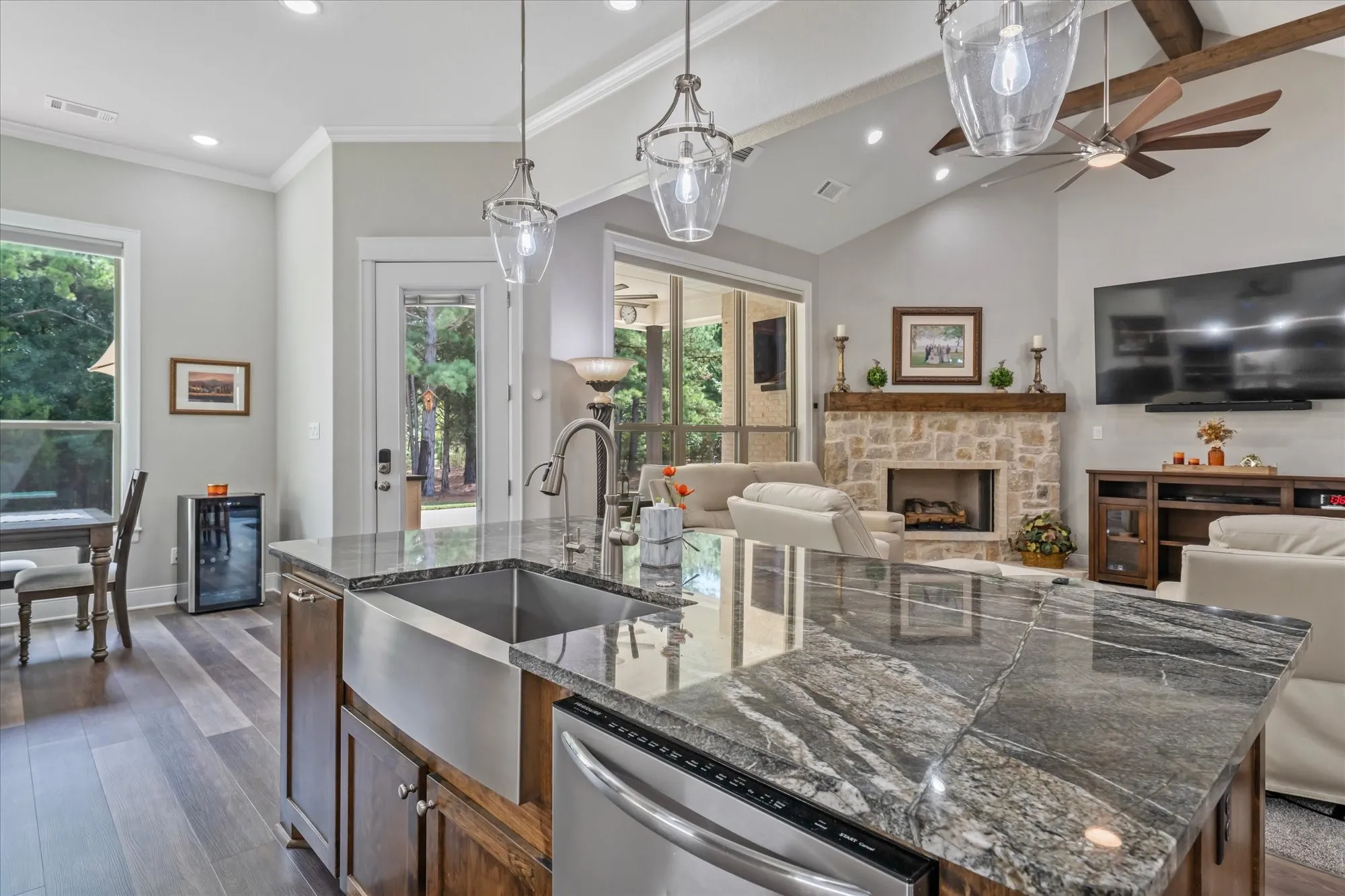 Vaulted ceiling with exposed wood beams, stone fireplace, wood-finish flooring, central island with an undermount stainless steel sink, and pendant lighting