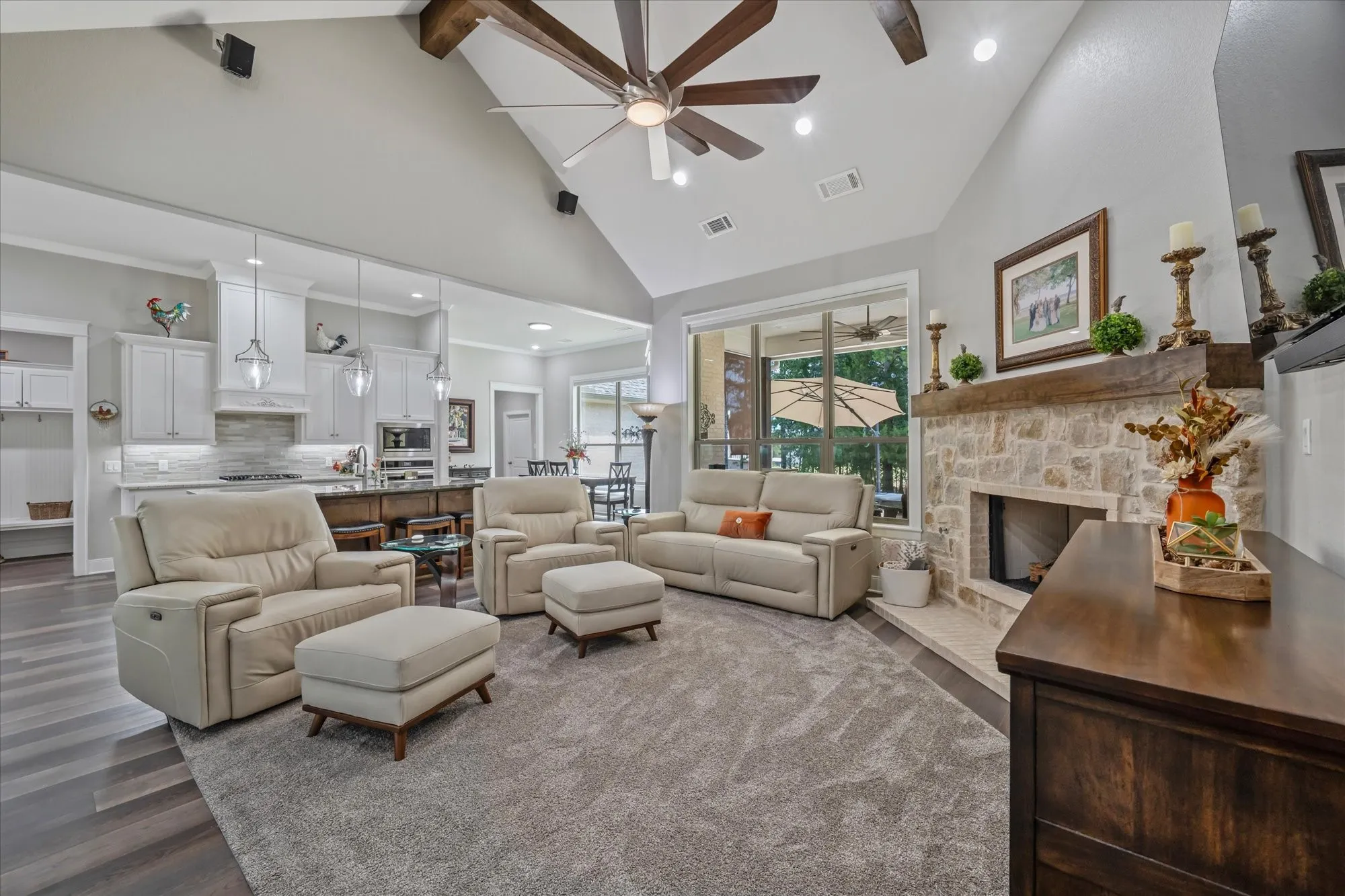 Vaulted ceiling with exposed wood beams, stone fireplace with a wood mantle, wood-finish flooring, white cabinetry, and pendant lighting