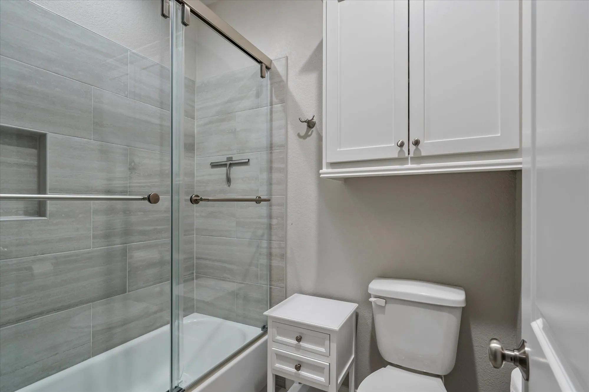 Bathtub with sliding glass shower door, gray tiled shower surround, recessed shower niche, and wall-mounted white cabinetry