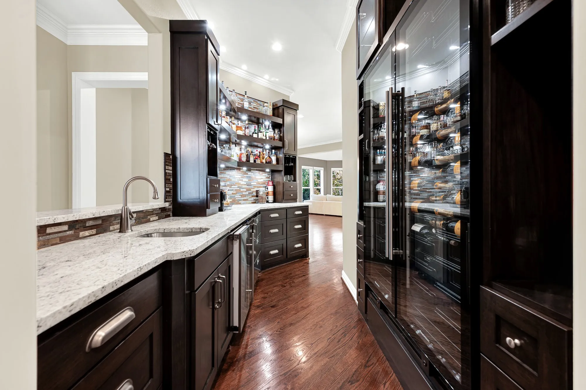 Floor-to-ceiling glass display fridges glowing with warm interior lighting anchor this custom passage. Pour a favorite vintage after work as soft light reflects off dark polished millwork