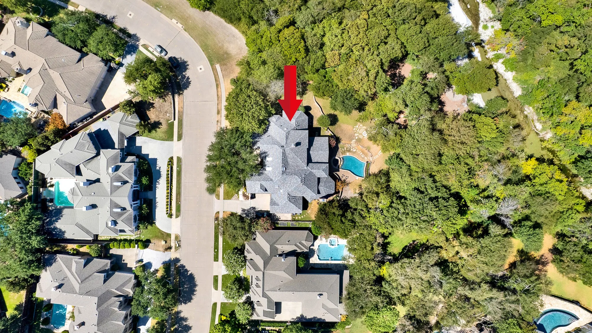 A top-down perspective showcases the intricate footprint nestled between a curving street and a winding creek bed. Start crisp mornings enjoying coffee as sunlight catches the sparkling blue pool tucked behind the trees.