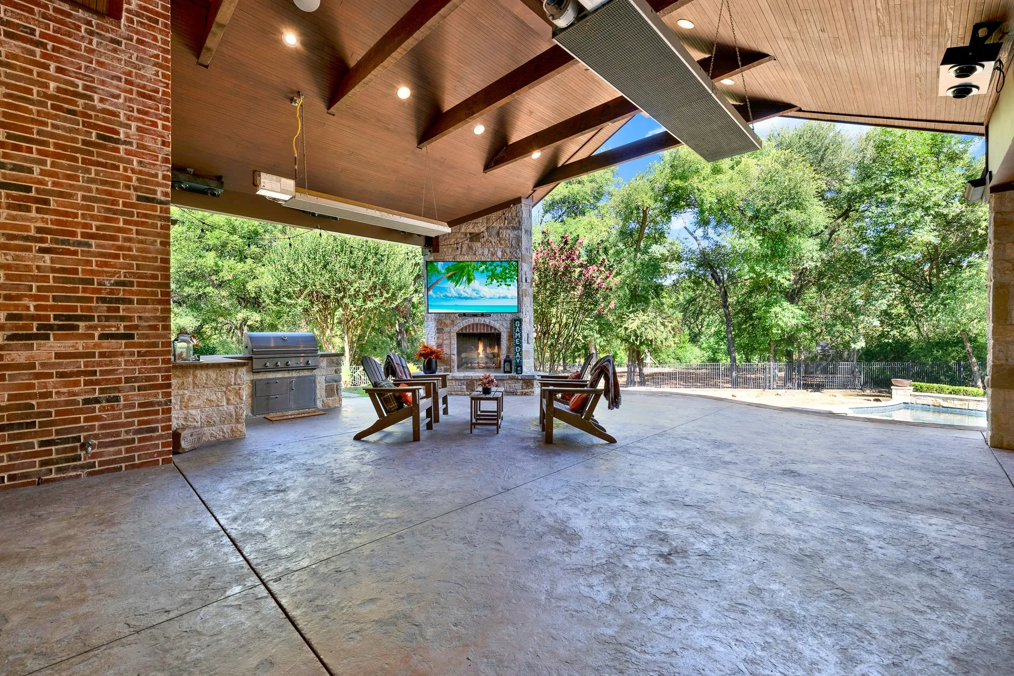 A stacked-stone outdoor fireplace hearth stands beneath exposed wood ceiling beams, framing sweeping views of the lush yard. Warm autumn evenings draw family outside to unwind by a crackling fire as dusk settles.