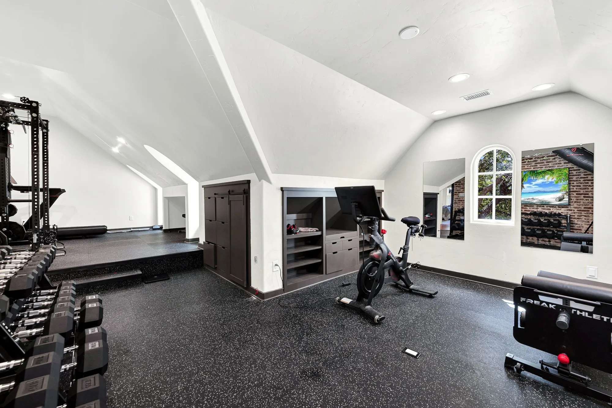 Angled rooflines create cozy architectural zones, while a charming arched window draws bright sunlight across the dark, resilient flooring. Start your day with an invigorating sunrise workout in this naturally lit retreat.