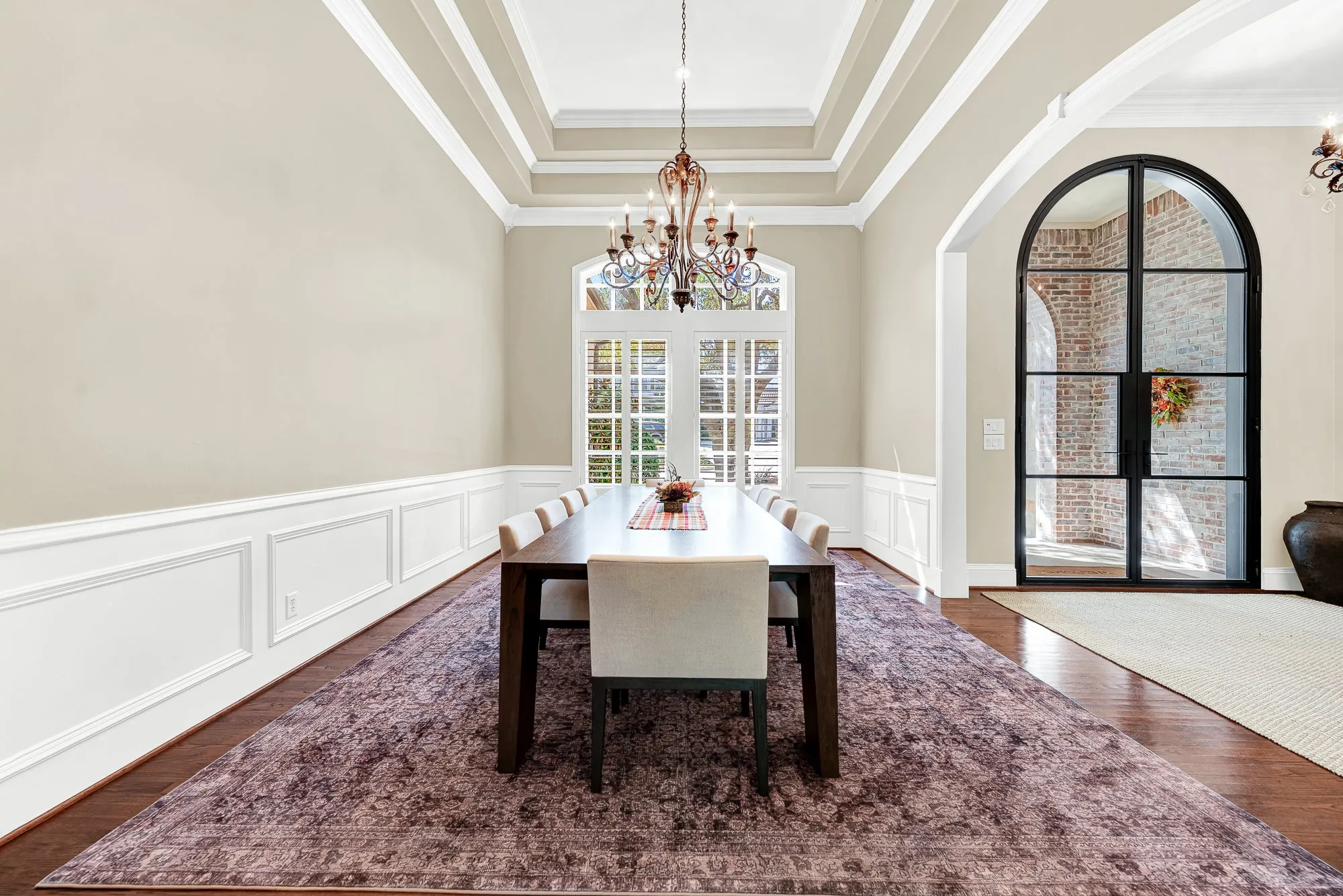 Layered tray ceilings and crisp wainscoting add tailored depth beneath a classic, glass-framed double door. You can almost hear the laughter during an intimate dinner party illuminated by soft chandelier light.