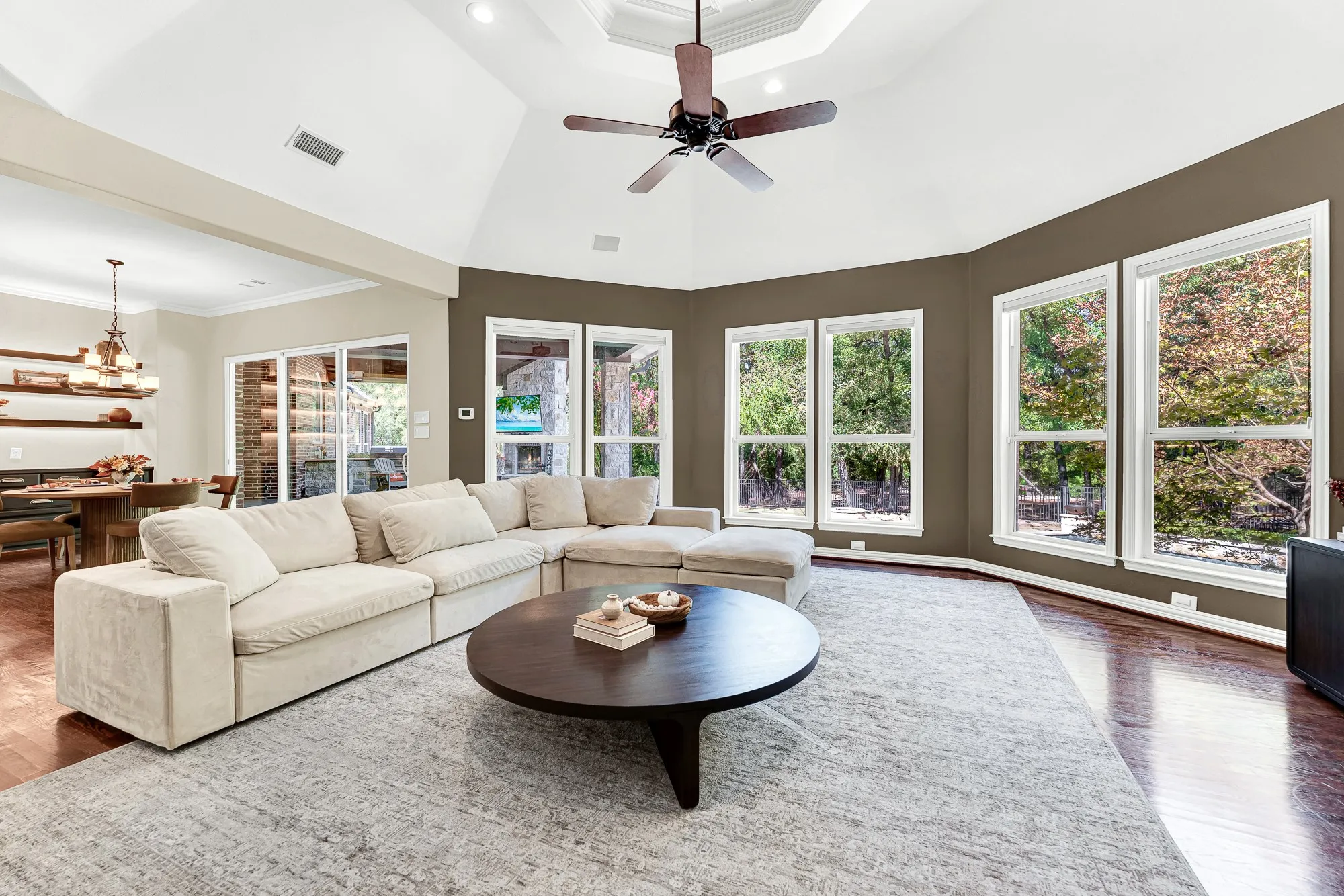 Panoramic bay-style windows frame leafy outdoor views, filling the high-ceilinged room with balanced daylight. Imagine hosting weekend movie nights bathed in the soft glow of dusk outside