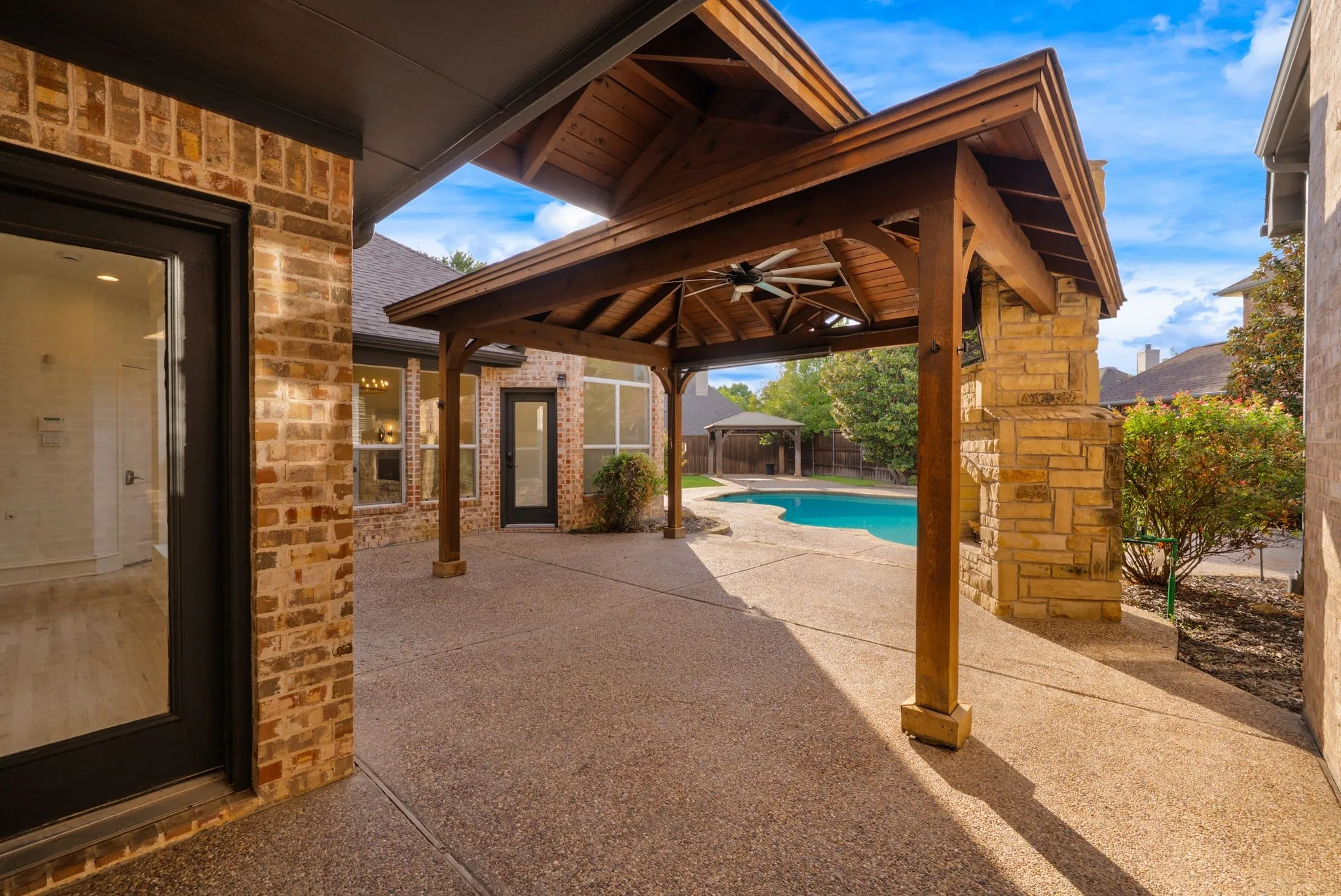 Expansive patio featuring exposed aggregate concrete, a wooden pergola with a ceiling fan, a stone-clad pillar, and a swimming pool