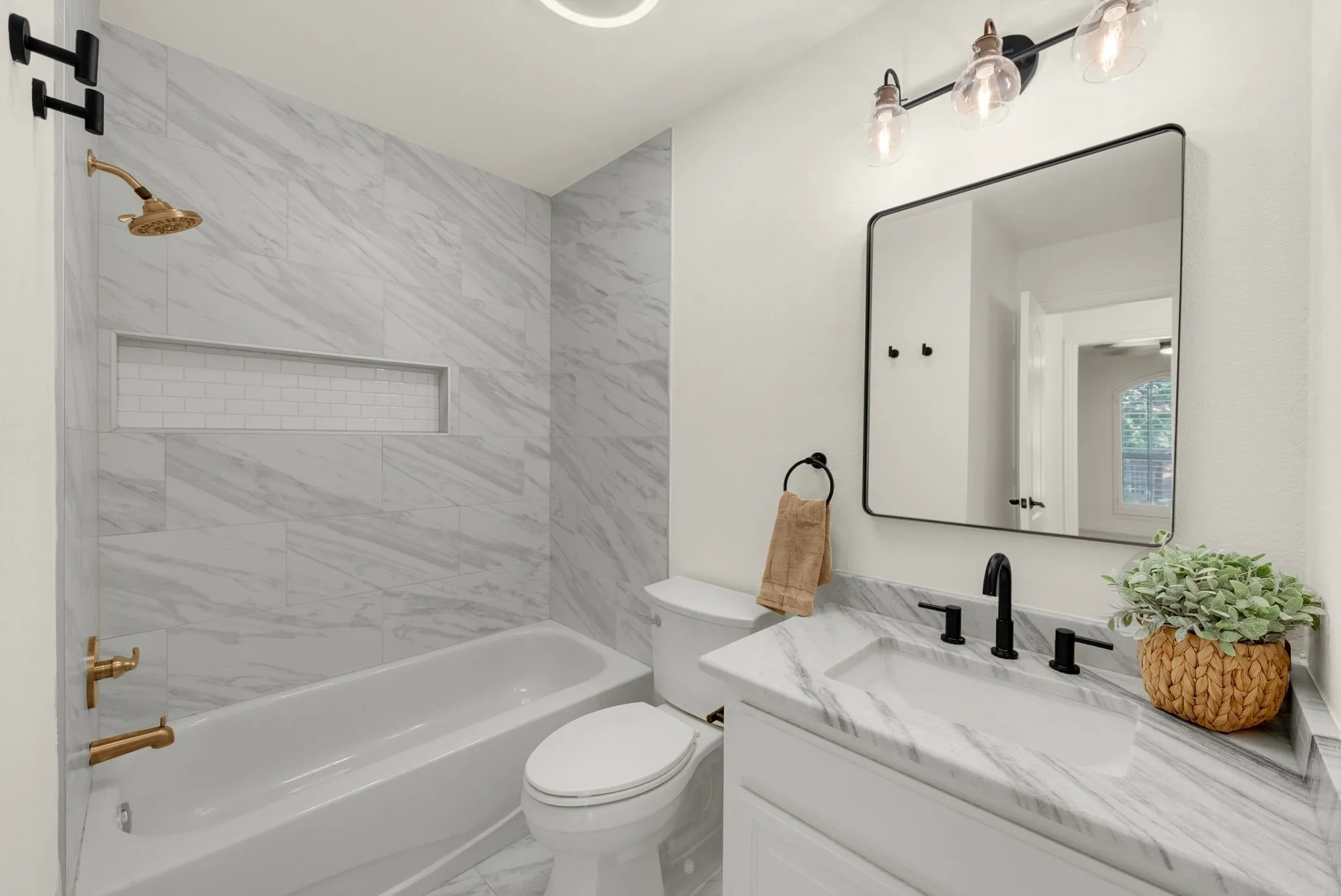White bathtub and toilet with brass fixtures, a built-in shower niche, and a vanity featuring a white rectangular sink, stone-finish countertop, and matte black faucet