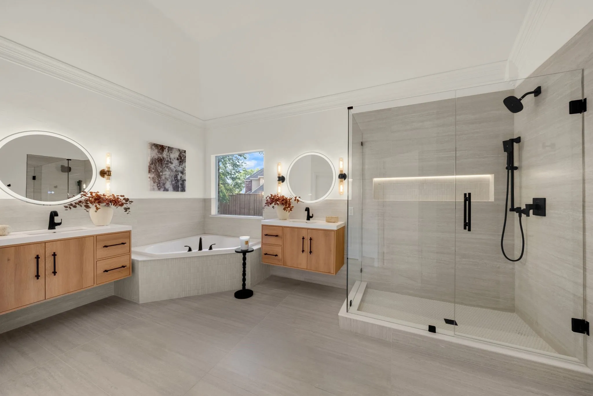 Spa-inspired primary bathroom featuring dual wood-finish vanities with integrated sinks, illuminated round mirrors, and matte black hardware
