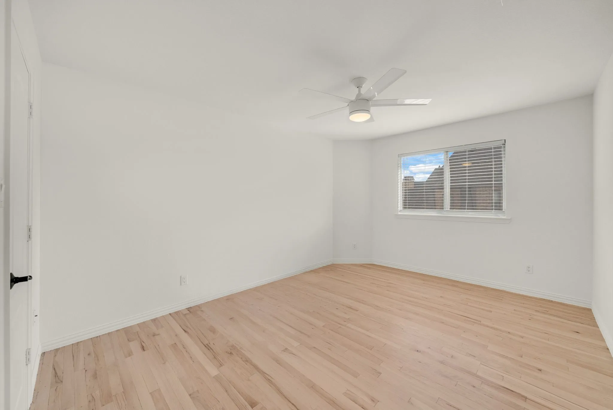 Light wood-finish flooring, white walls, ceiling fan with integrated lighting, and a window with blinds