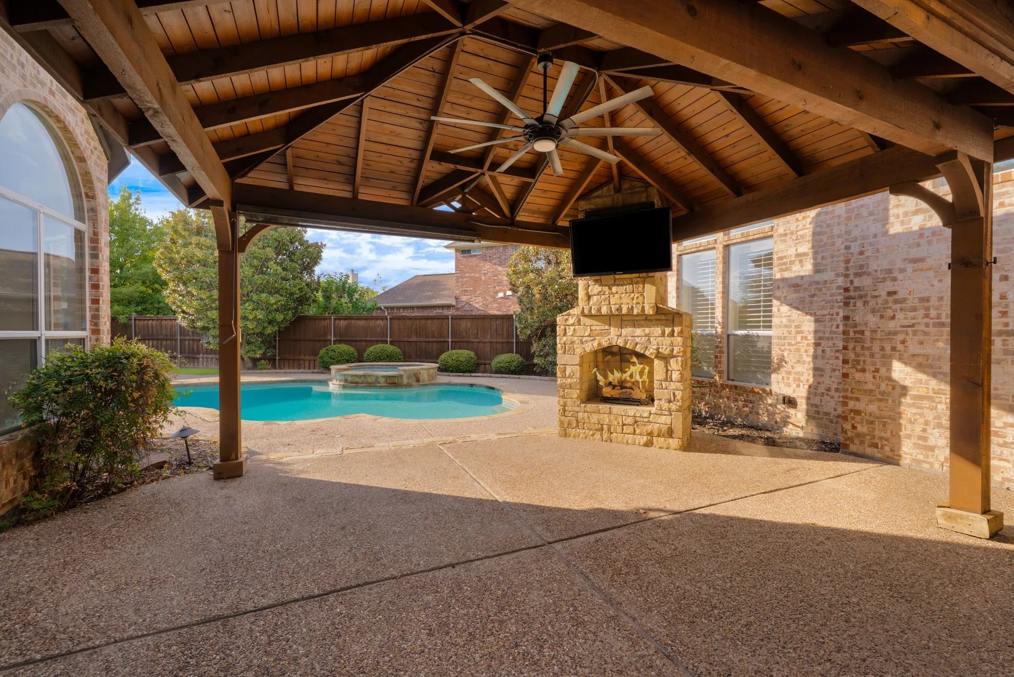 Expansive covered patio with exposed wood-beam ceiling and ceiling fan, anchoring a resort-style backyard featuring an in-ground swimming pool with surrounding concrete decking, mature landscaping, and a brick fireplace with chimney