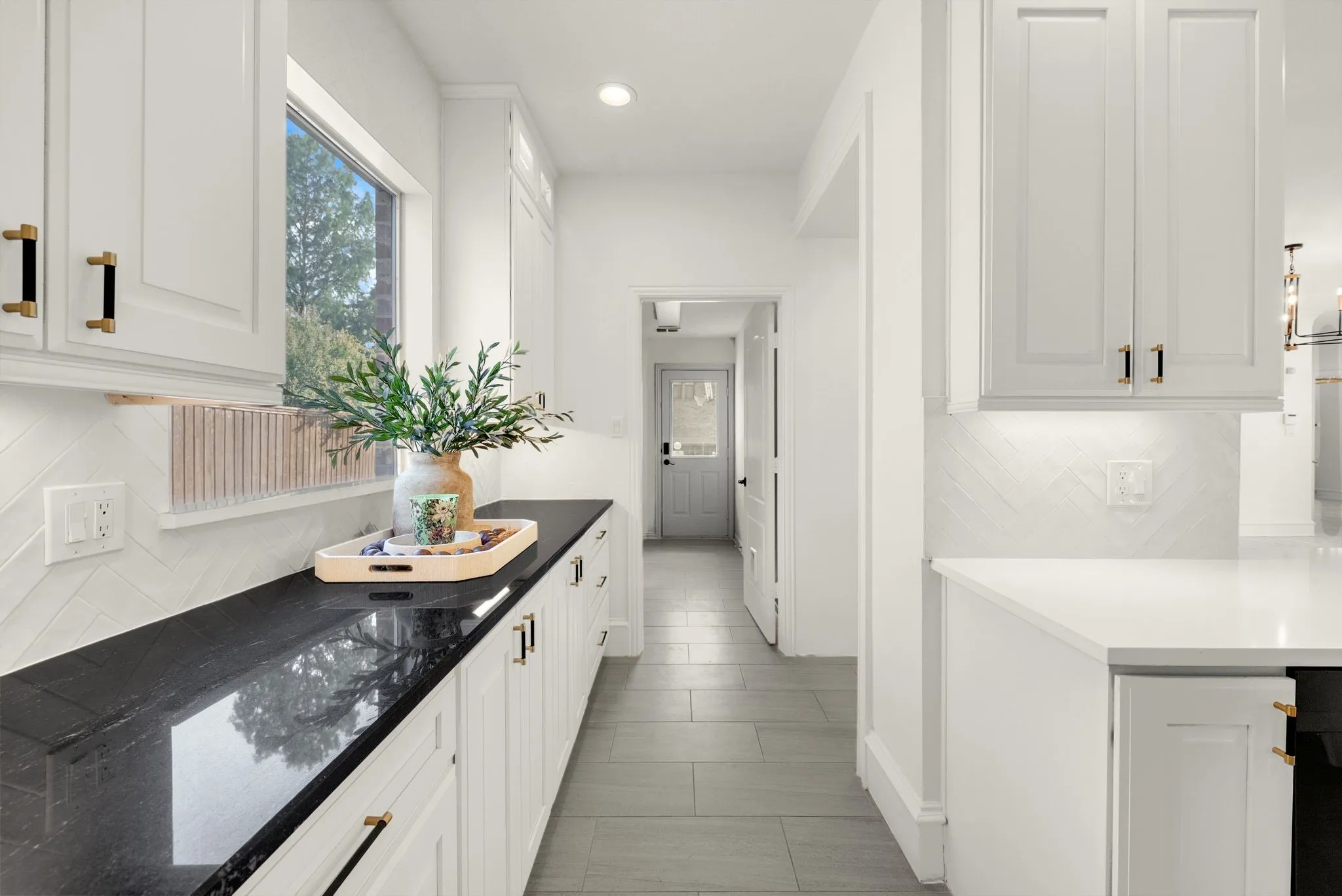 White shaker cabinetry with gold and black hardware, dark countertops, a window, white herringbone tile backsplash, and light-toned flooring