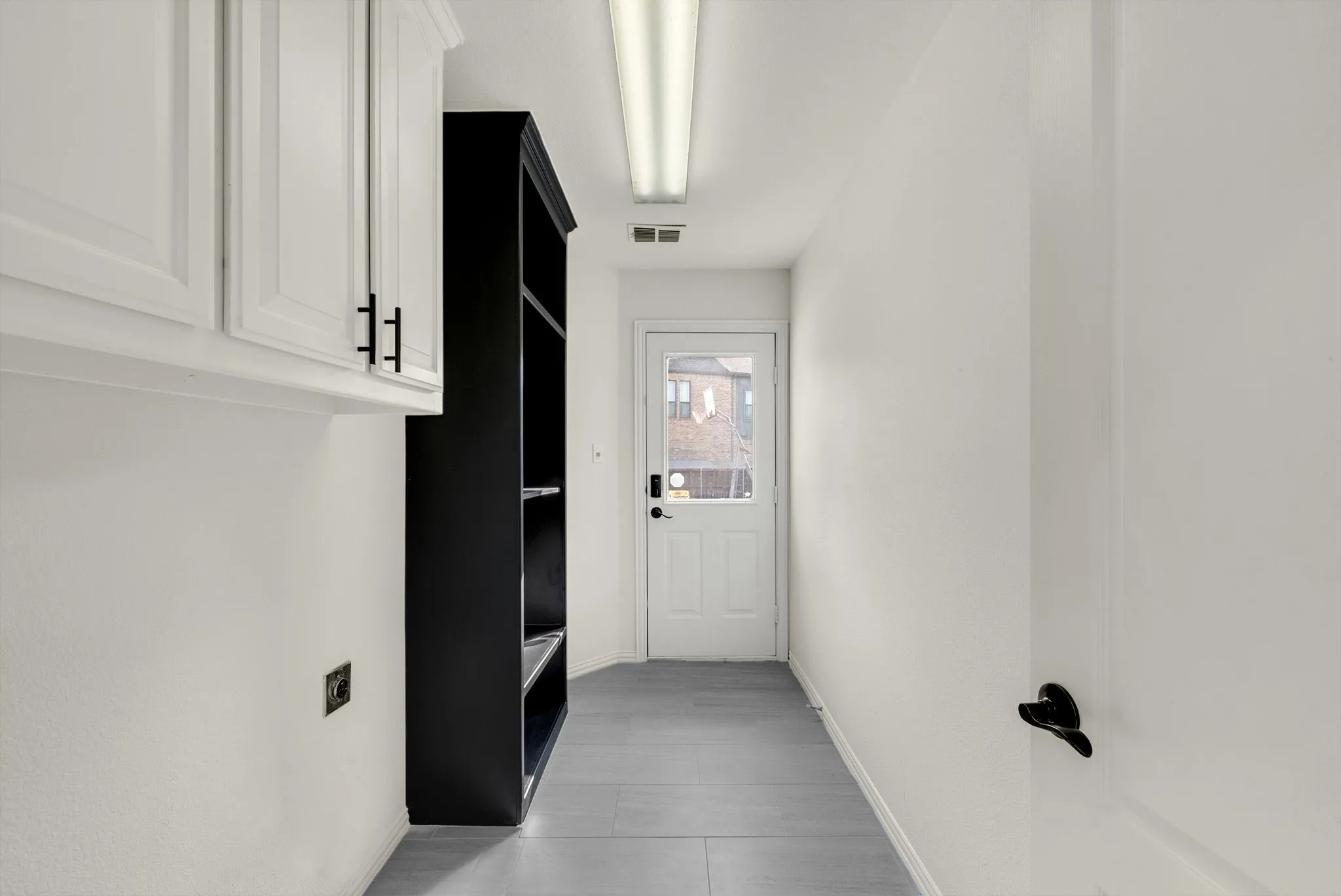 White upper cabinetry with black hardware, built-in black shelving, a white door with a glass panel, and gray tile flooring