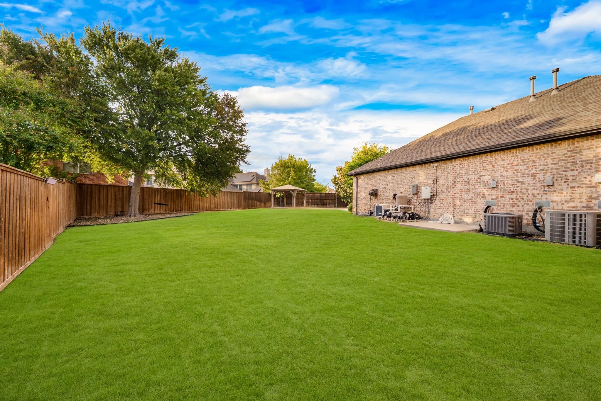 Expansive manicured lawn with mature trees and established landscaping, backing onto a brick-constructed residence featuring a patio area and detached outbuilding