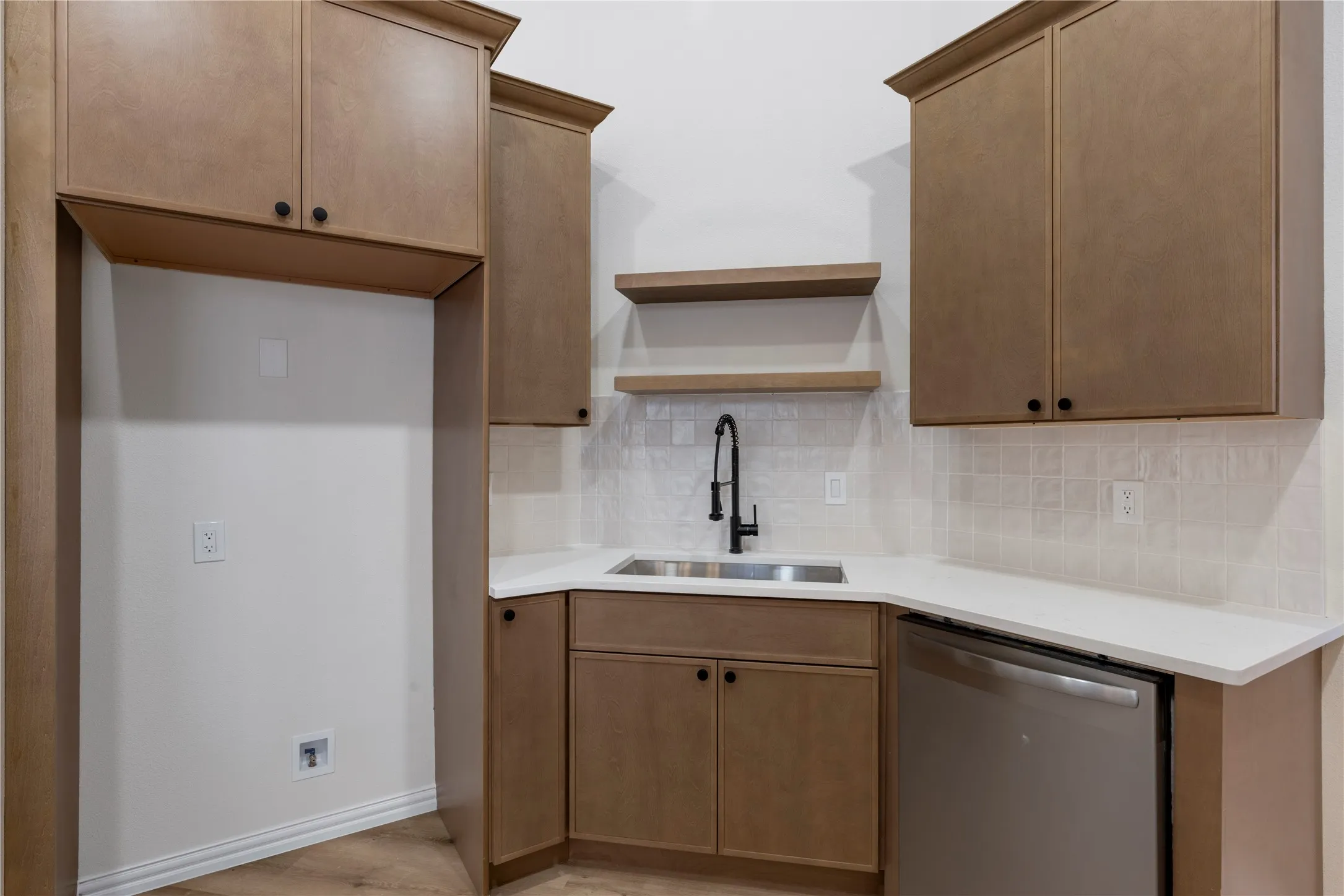 Corner sink with a matte black faucet, stainless steel dishwasher, light-toned cabinetry, two floating shelves, and white countertops
