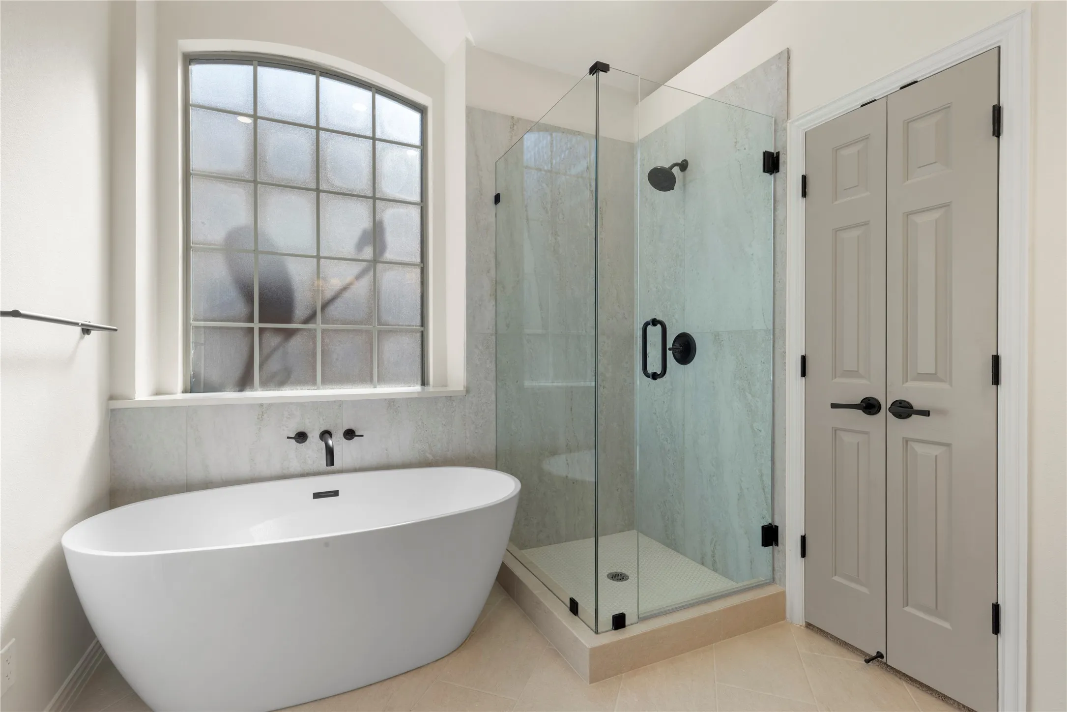 Freestanding soaking tub with wall-mounted matte black faucet, glass-enclosed shower with matte black fixtures, and arched window with frosted glass