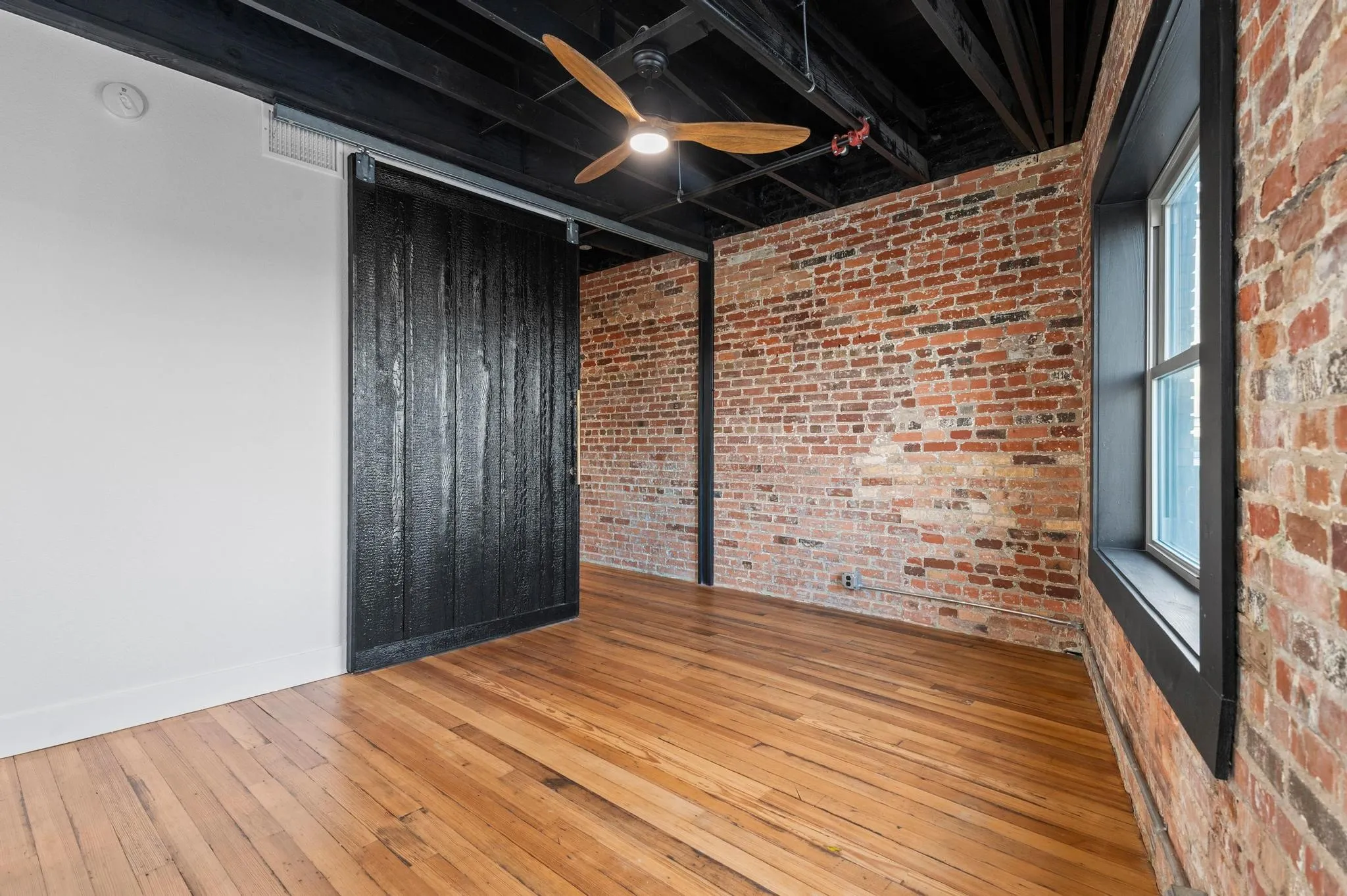 Exposed brick walls, wood-finish flooring, industrial ceiling with exposed ductwork, barn door, and a window with dark trim