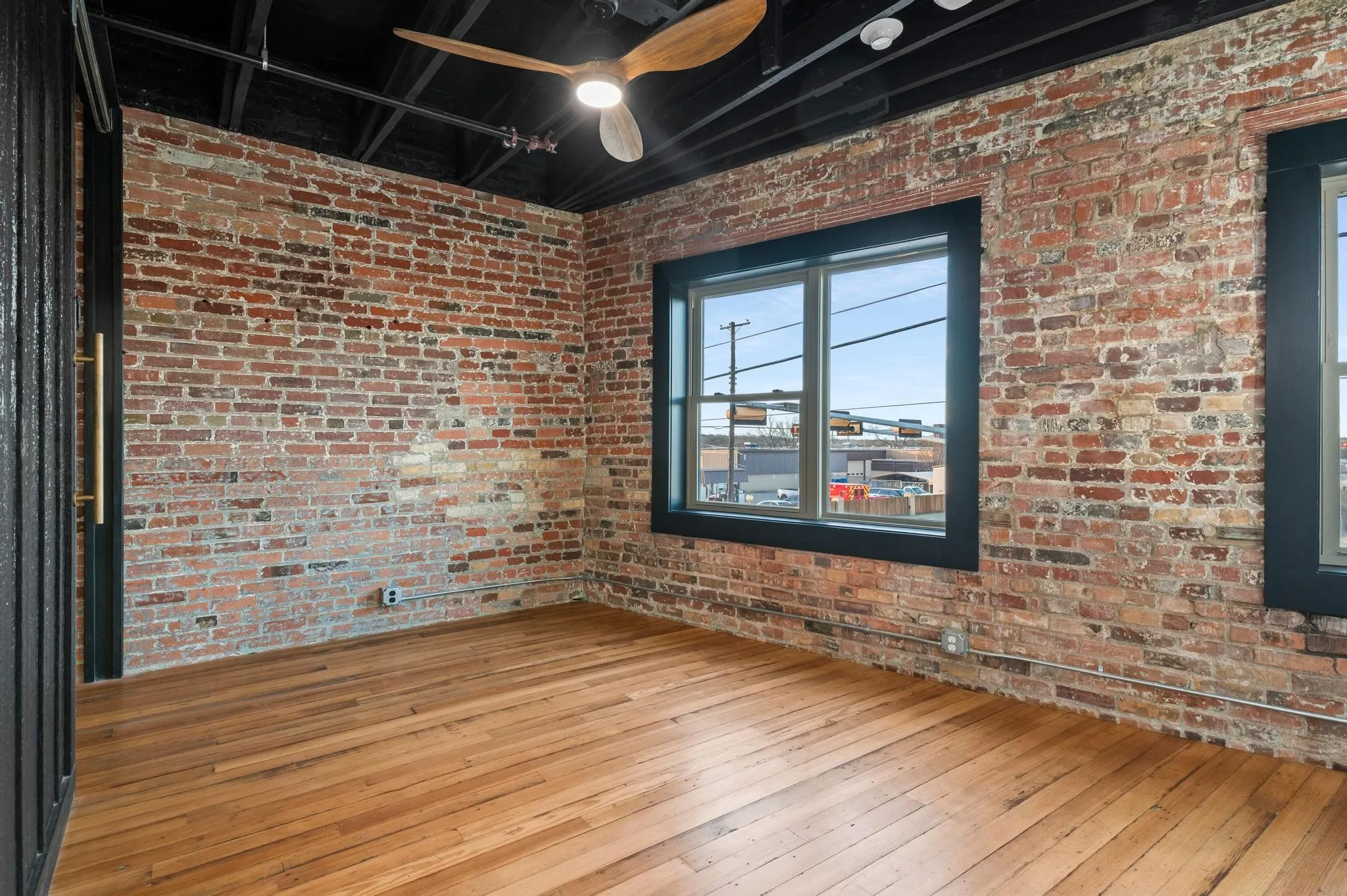 Exposed brick walls, wood-finish flooring, two large windows with black trim, black ceiling with exposed ductwork, and a ceiling fan