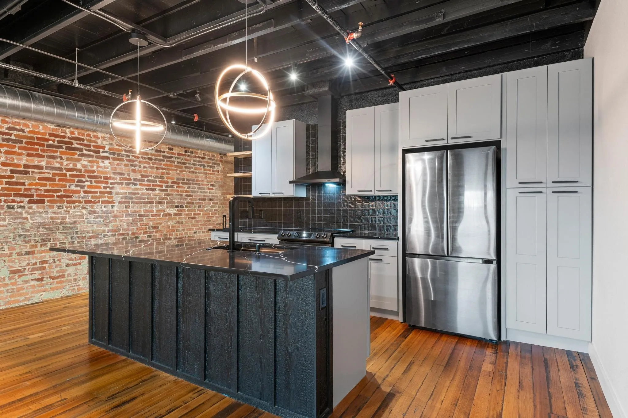 Exposed brick wall, hardwood flooring, modern pendant lighting, stainless steel refrigerator, and light-toned cabinetry