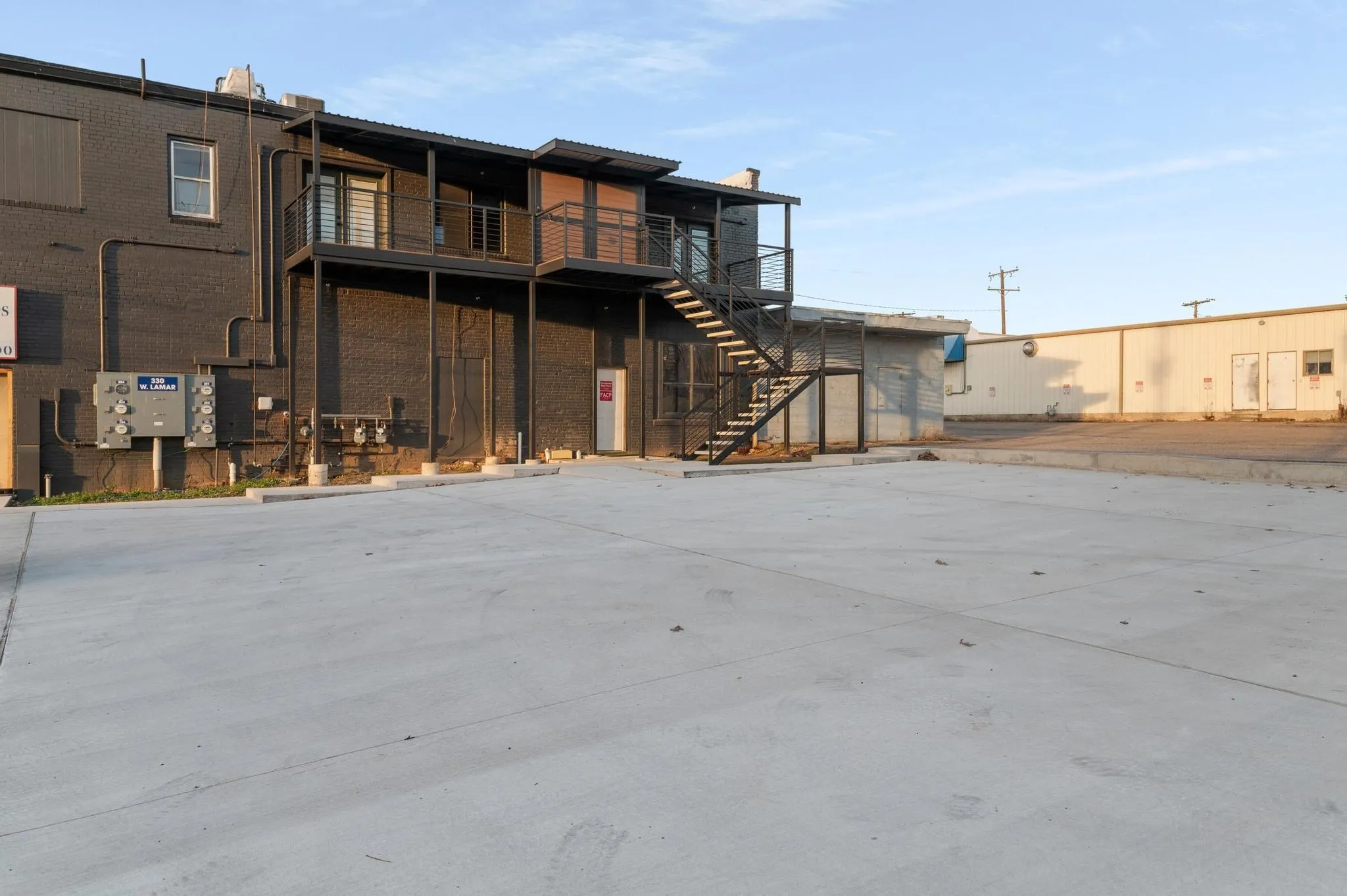 Paved concrete parking area with a two-story building featuring a dark brick exterior, an exterior metal staircase, and a second-story balcony