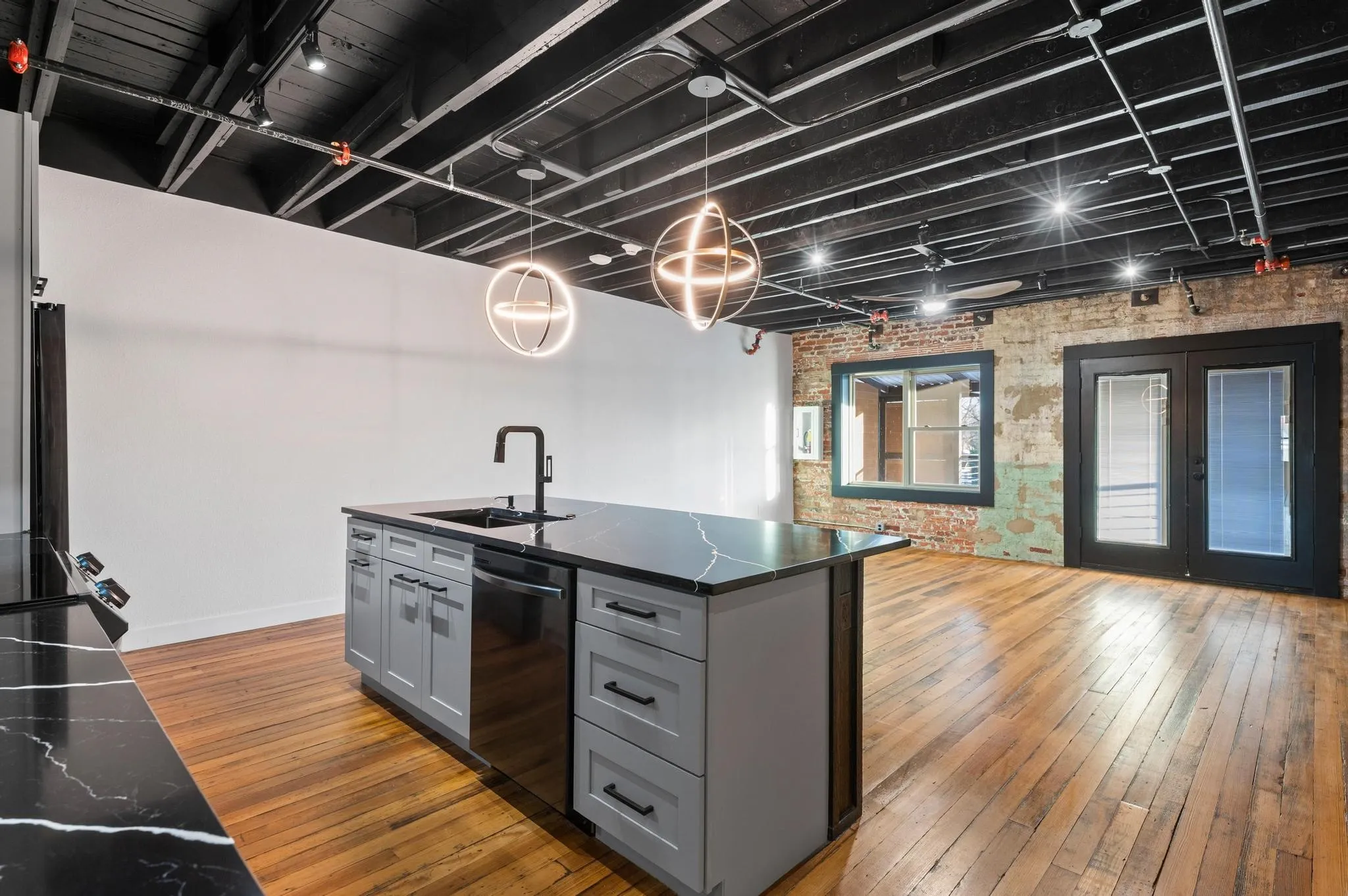 Exposed ceiling with track lighting, hardwood flooring, brick accent wall, kitchen island with dark countertops, and black French doors