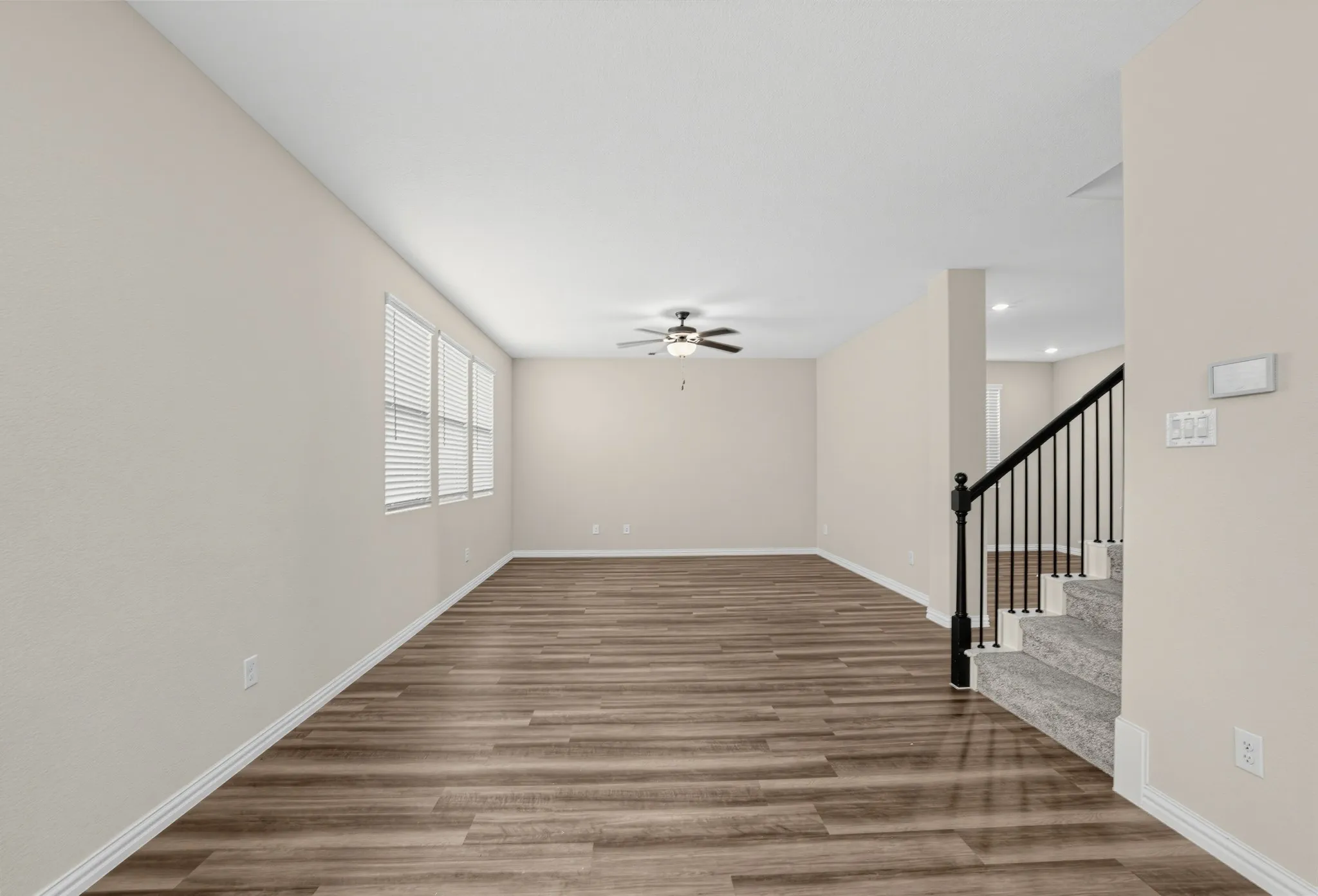 Spacious living area featuring wood-finish flooring, multiple windows with blinds, a ceiling fan, and a staircase with a wrought-iron railing