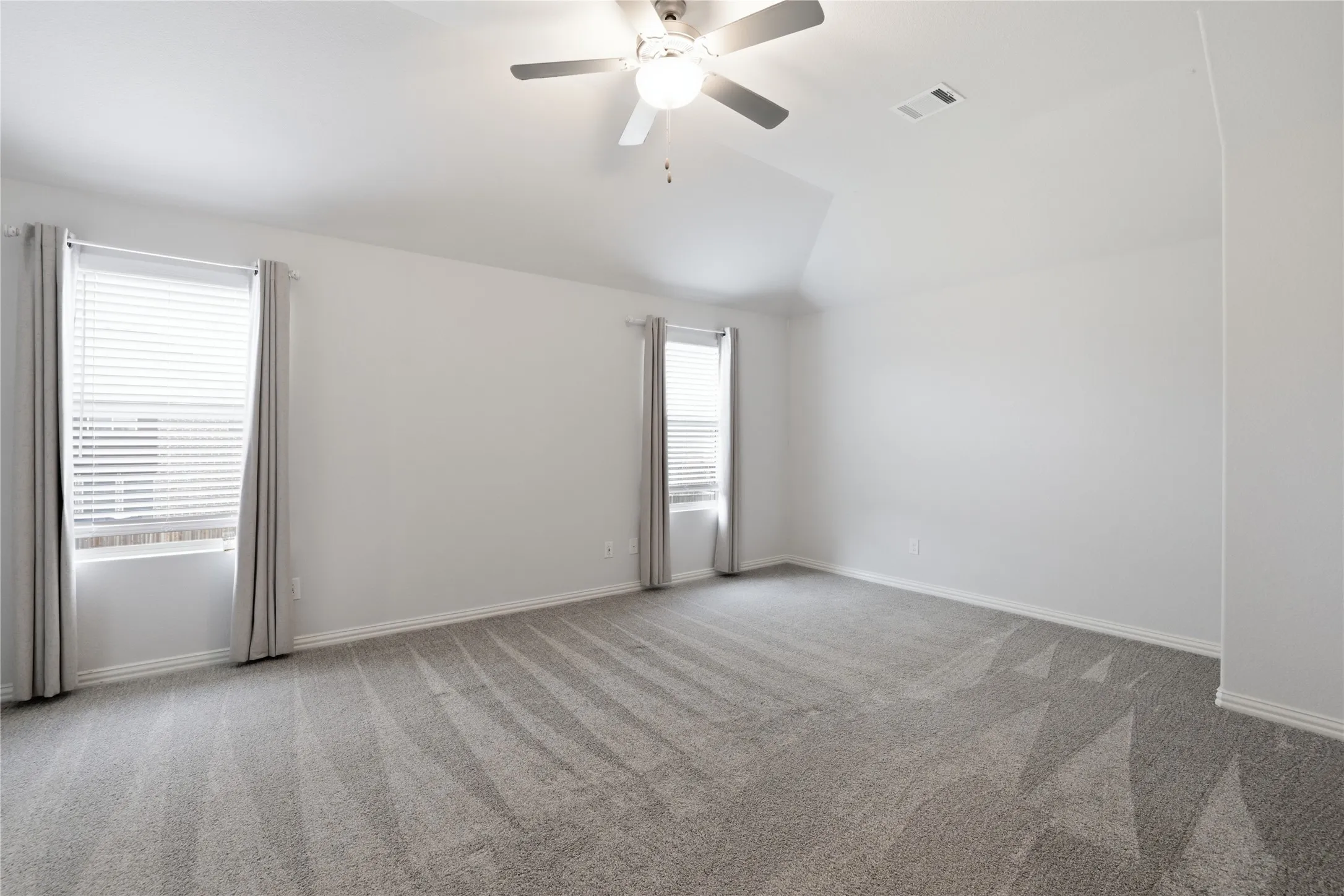 Spacious room featuring vaulted ceilings, a ceiling fan with integrated lighting, two windows with blinds, and neutral carpeting