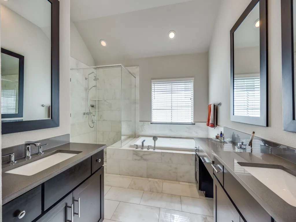 Dual vanities with integrated sinks and dark cabinetry, large soaking tub, glass-enclosed shower with tiled walls, natural light from a privacy window, and recessed lighting