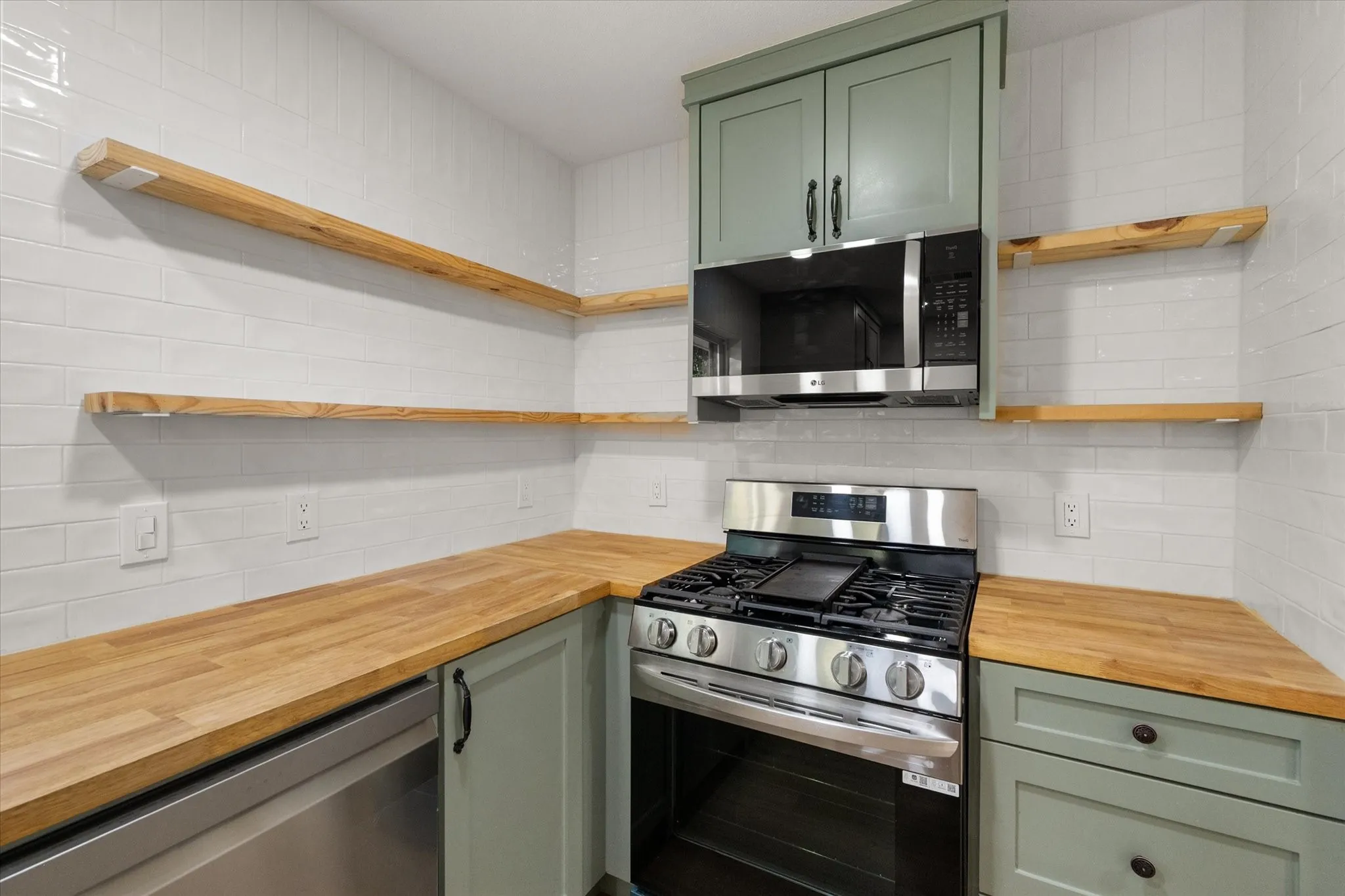 White subway tile backsplash, butcher style open shelving and counter tops, stainless steel range, stainless steel microwave and shaker style painted cabinetry with hardware.