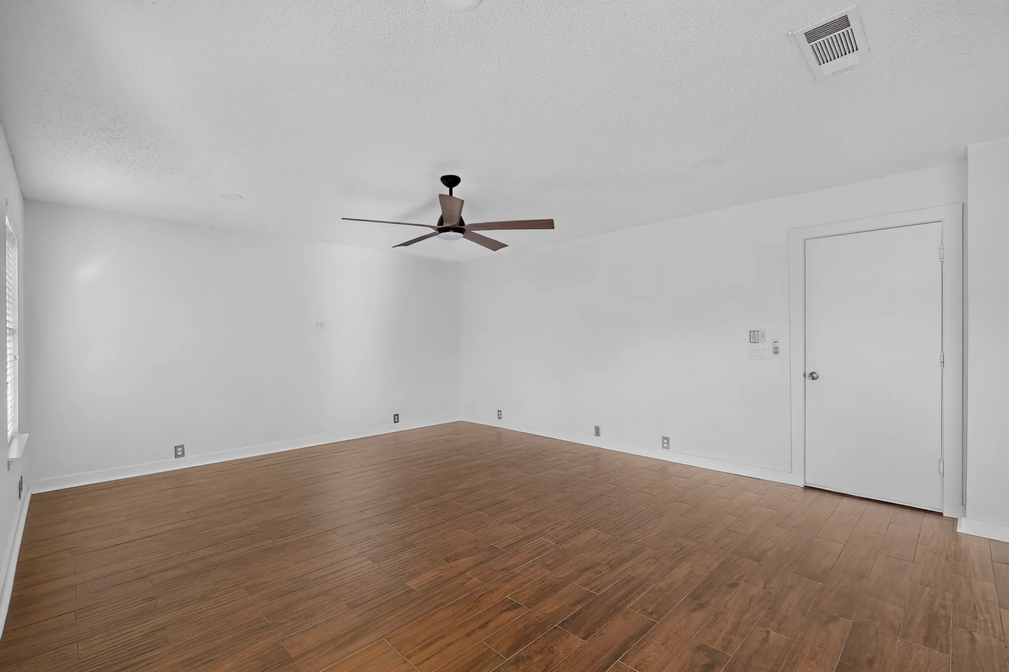 Large family room with windows looking the back yard with wood-finish flooring, crisp white walls, ceiling fan with integrated lighting.
