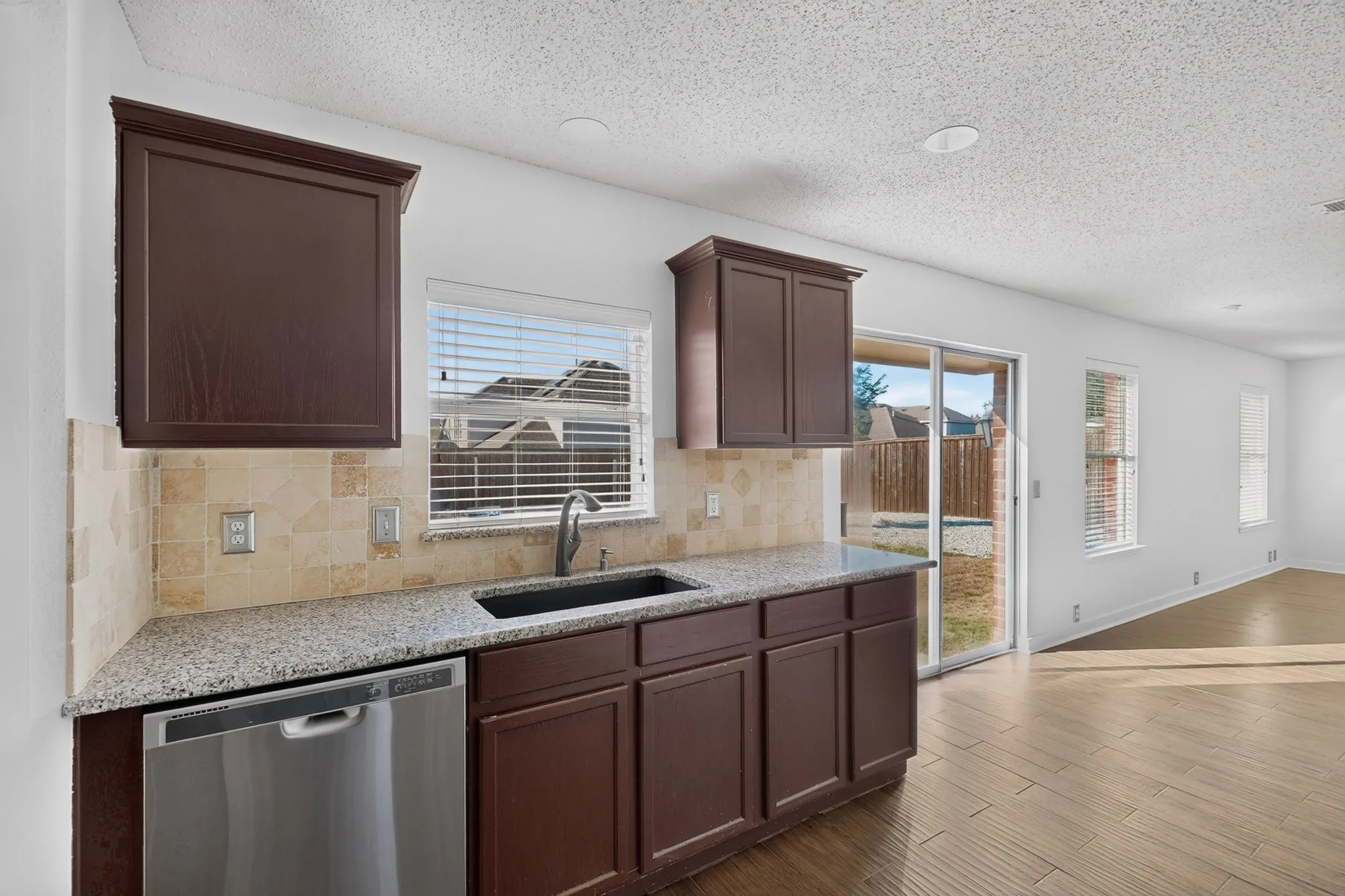 Kitchen featuring wood-finish cabinetry, granite countertop, tile backsplash, stainless steel dishwasher, and a black undermount sink