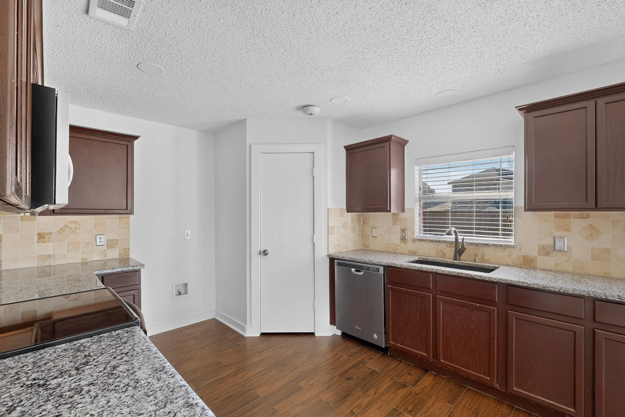 Kitchen featuring wood-finish flooring, dark wood cabinetry, walk-in pantry, light-toned backsplash and granite countertops.