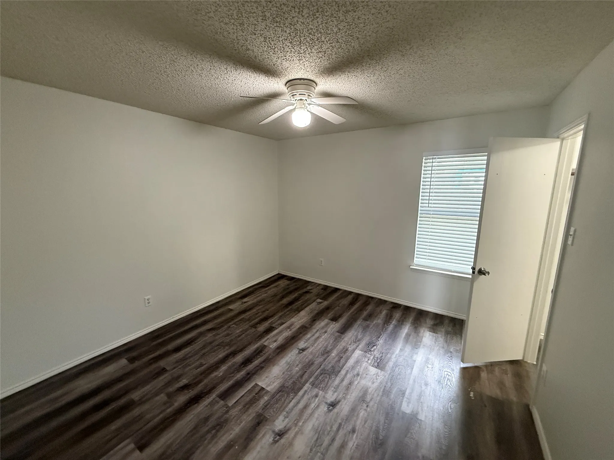 Wood-finish flooring, single window with horizontal blinds, overhead ceiling fan with lighting, and a white interior door
