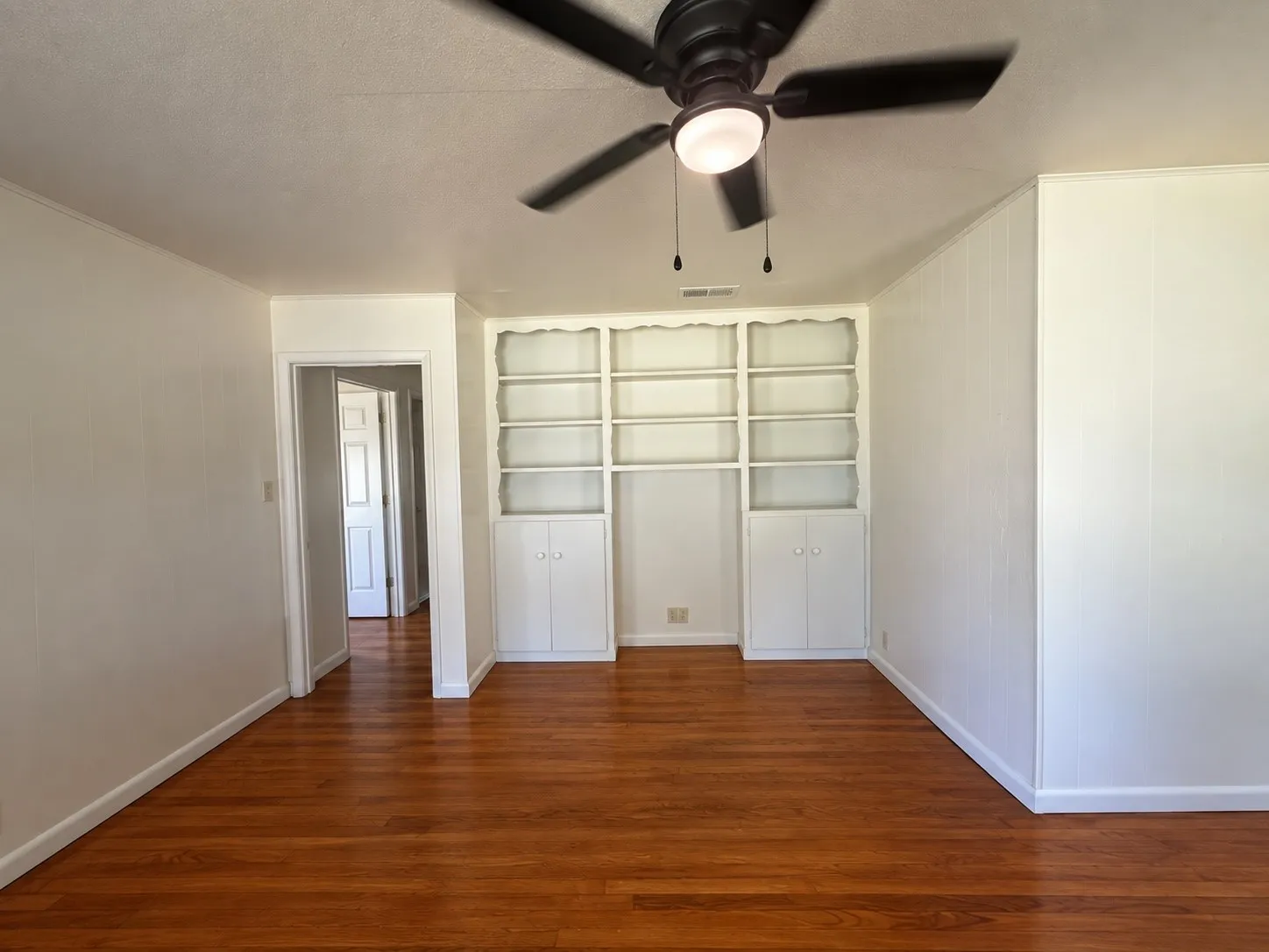Wood-finish flooring throughout, integrated built-in shelving, white wall paneling, and a ceiling fan with integrated lighting