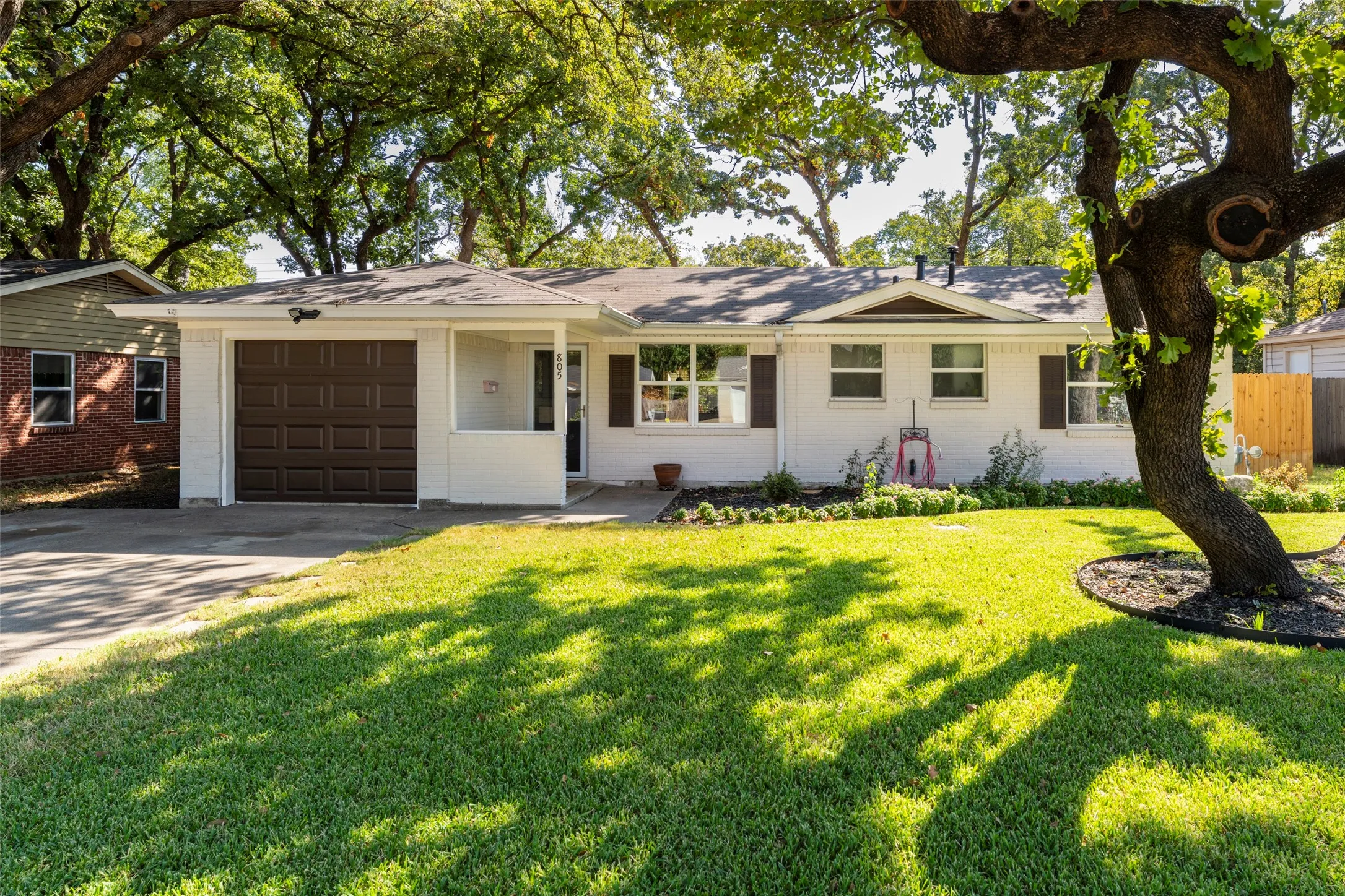 White brick exterior, dark brown garage door, front porch, dark brown window shutters, mature trees
