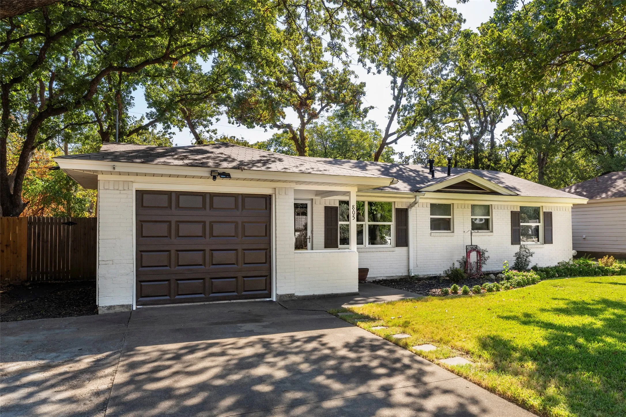 White brick exterior with dark shutters, an attached garage, a concrete driveway with stone accents, and mature trees
