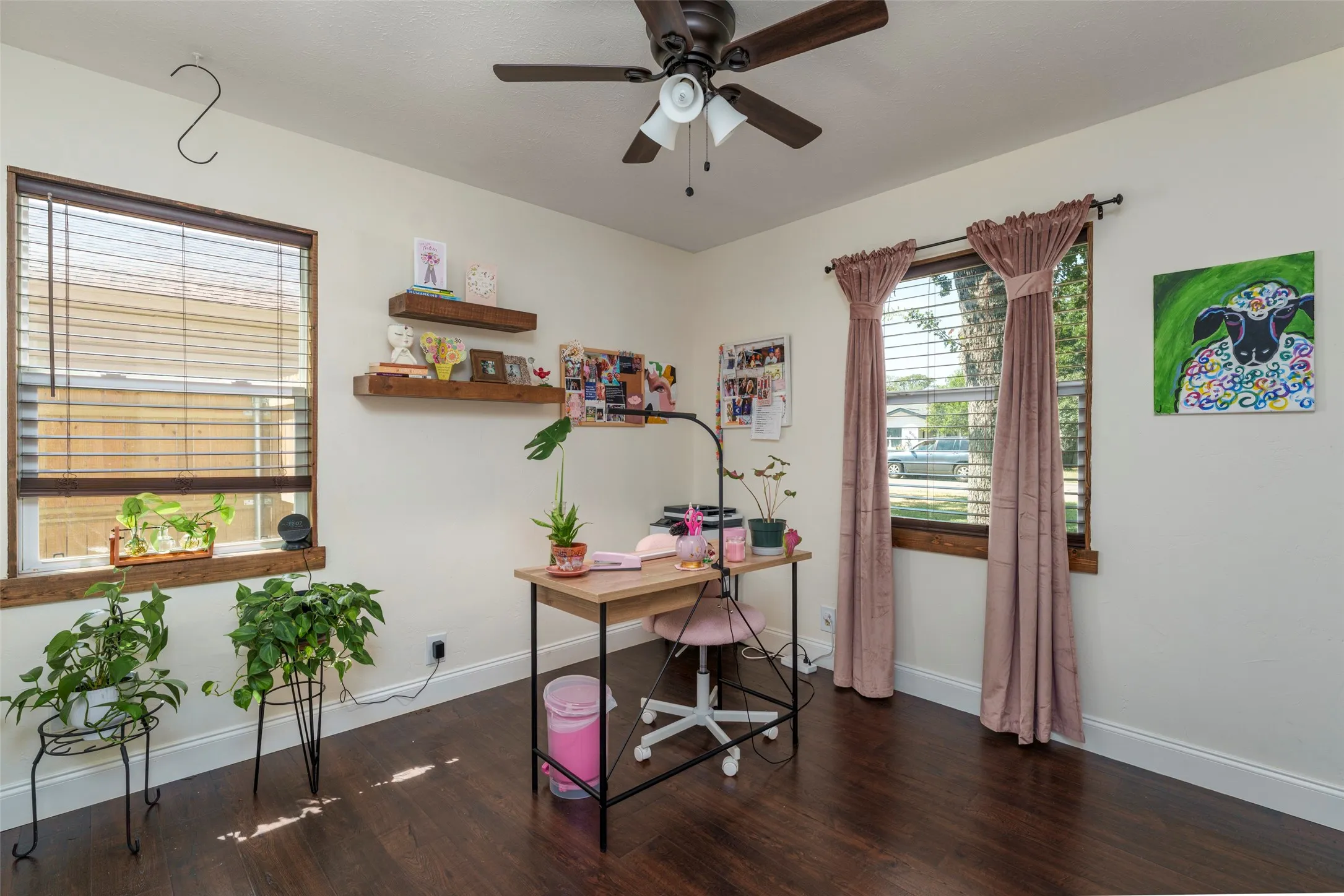 Dark wood-finish flooring, two windows with wood trim, ceiling fan with integrated lighting, and two floating wall shelves