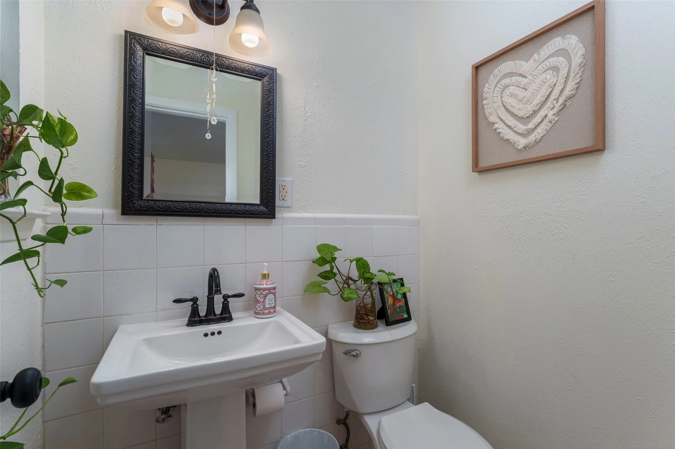 Pedestal sink with dark-finish faucet, framed mirror, wall-mounted light fixture, and white tile wainscoting