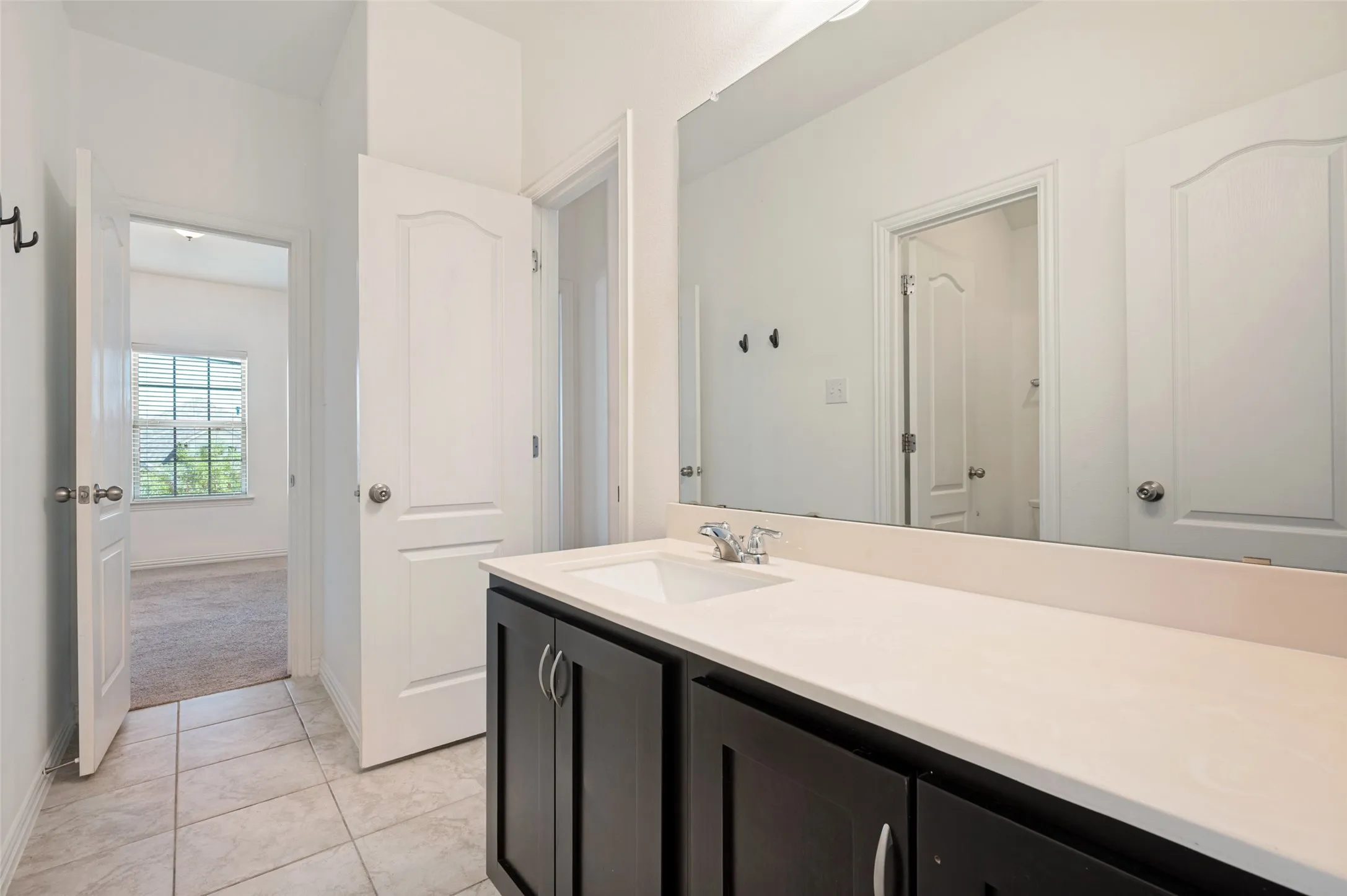 Vanity with a white countertop and integrated sink, chrome faucet, expansive wall mirror, dark wood-finish cabinetry, and tiled flooring