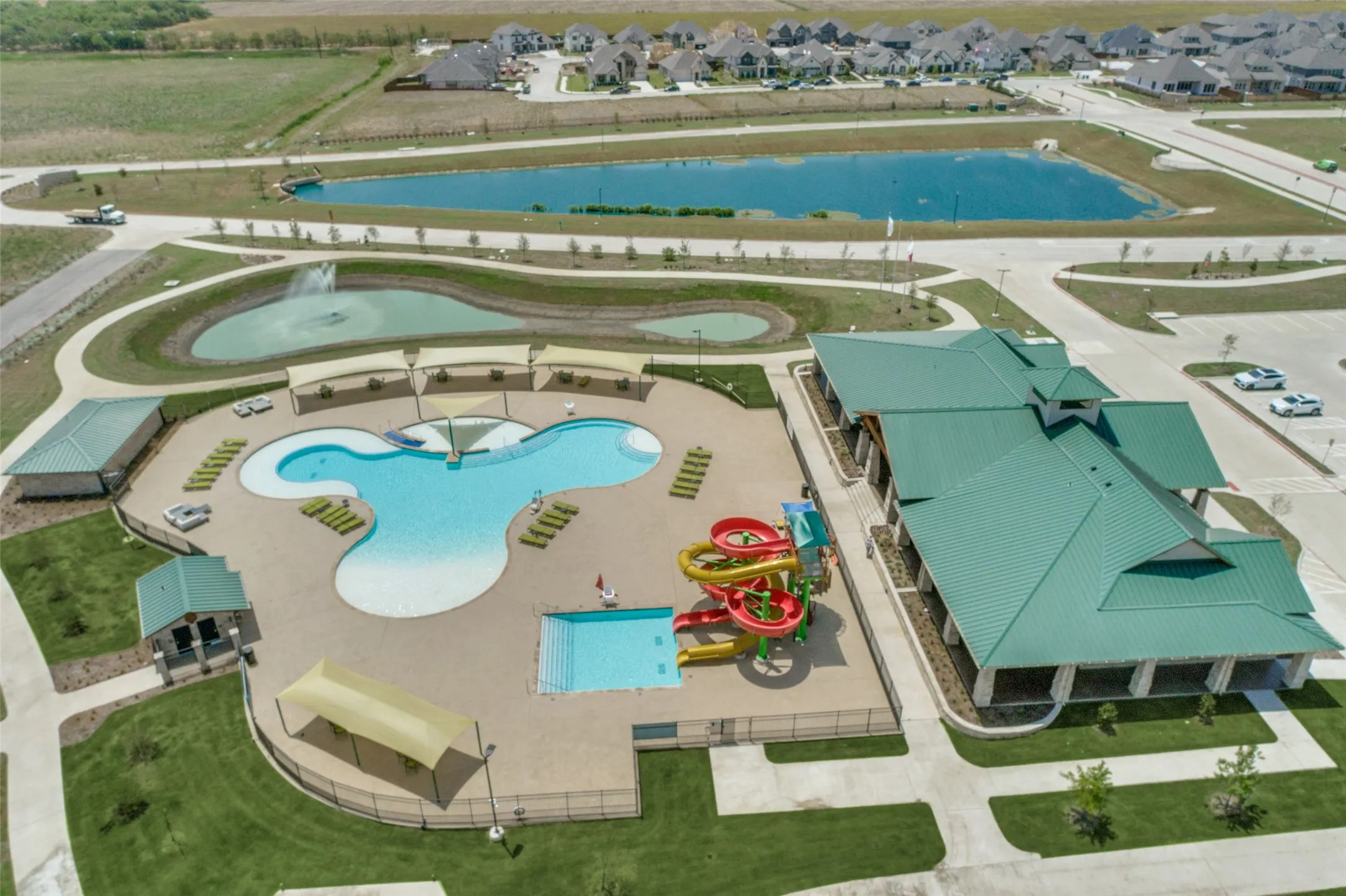 Community amenity center featuring a multi-level pool with water slides, extensive pool deck, shaded lounge areas, and adjacent building with covered patio