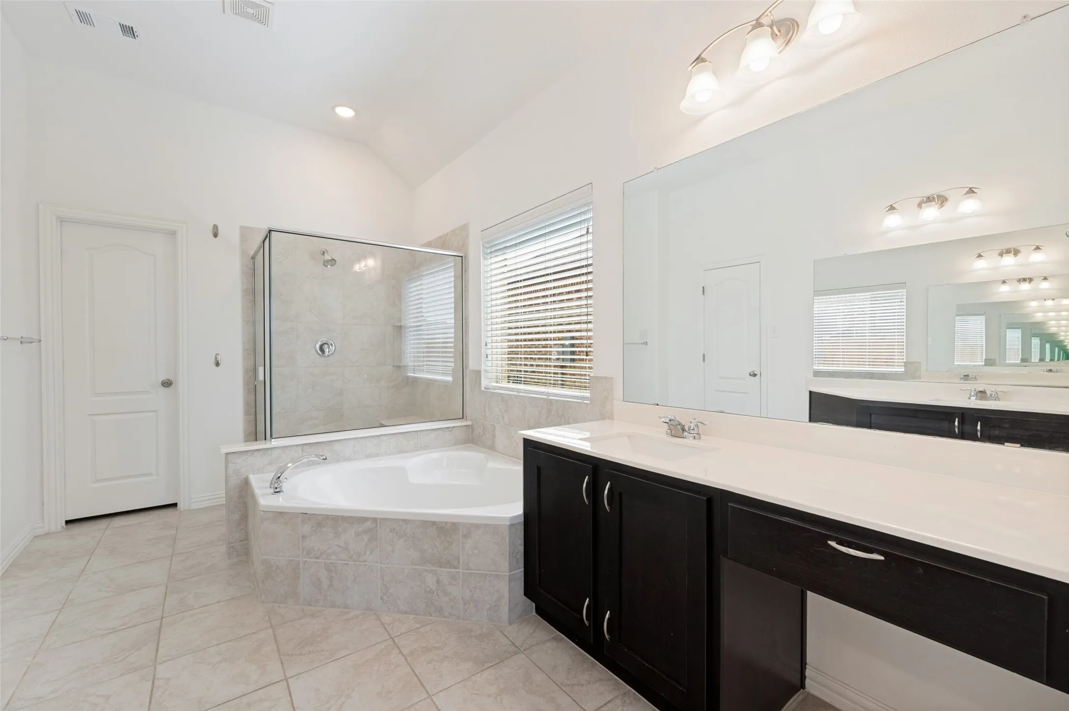 Tiled flooring, corner soaking tub, glass-enclosed shower, dark wood-finish vanity with white countertop, and expansive wall mirror