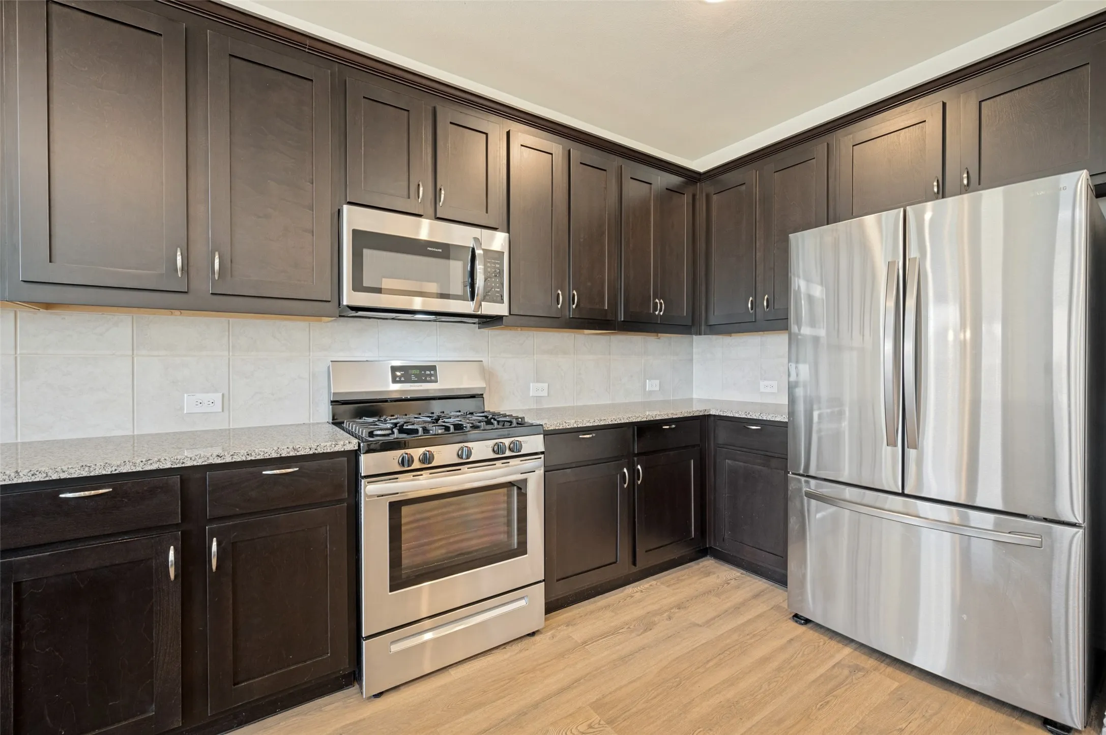 Dark-stained cabinetry, light-toned countertops, stainless steel appliances, tile backsplash, and wood-finish flooring