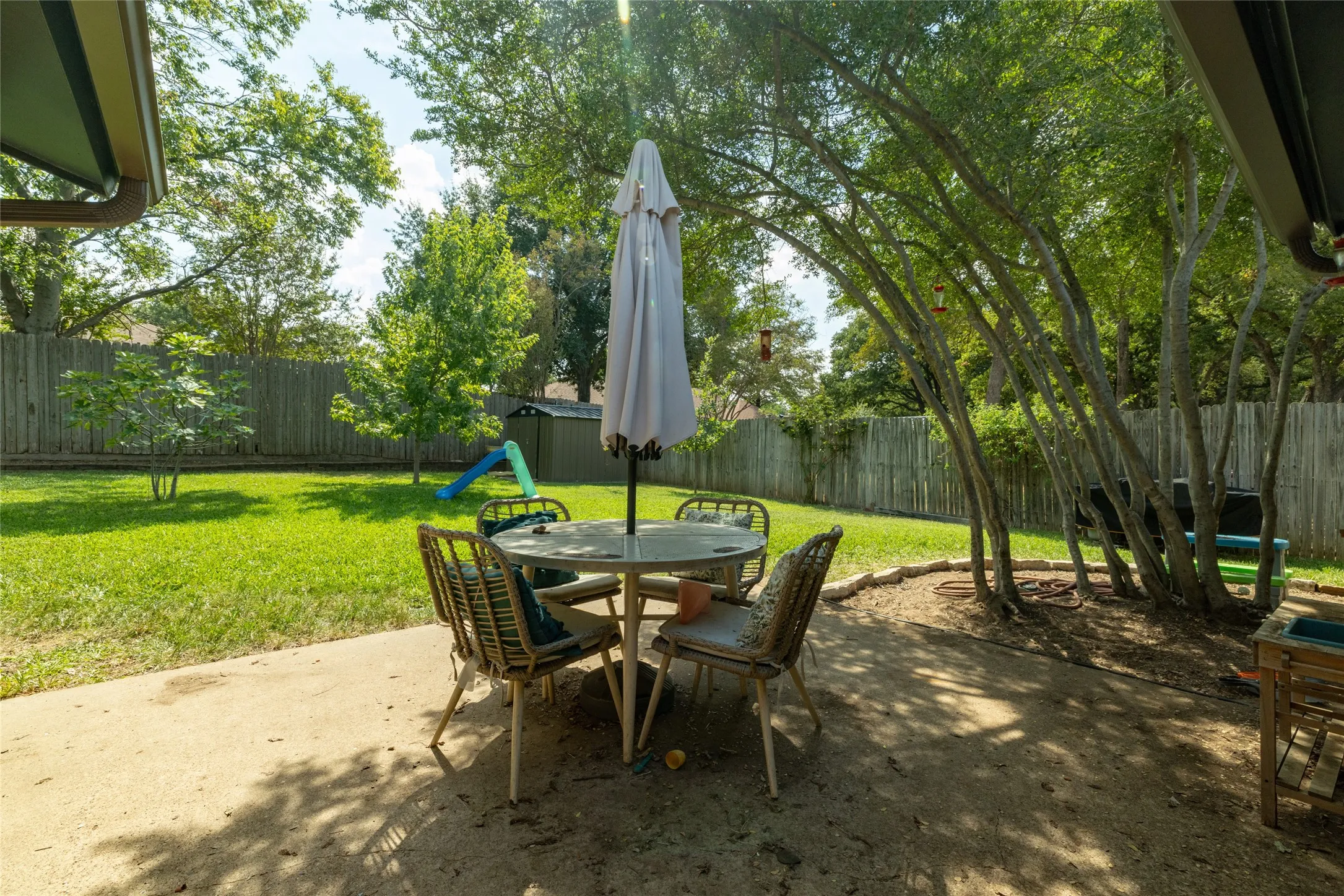 Expansive concrete patio with mature trees, a privacy fence, and a storage shed