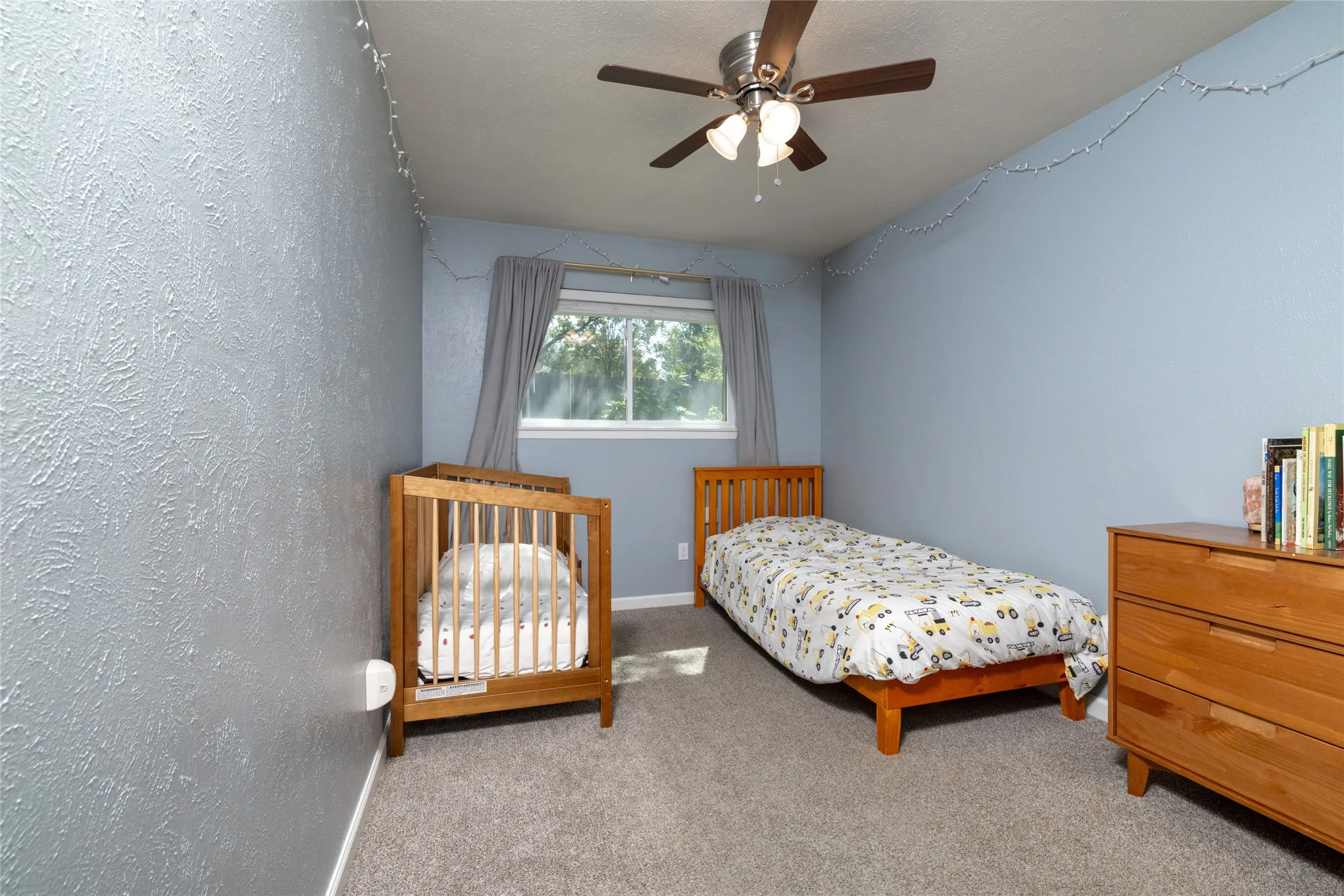 Textured wall finishes with contrasting smooth walls, a ceiling fan with integrated lighting, a window with drapery, and a gray carpeted floor
