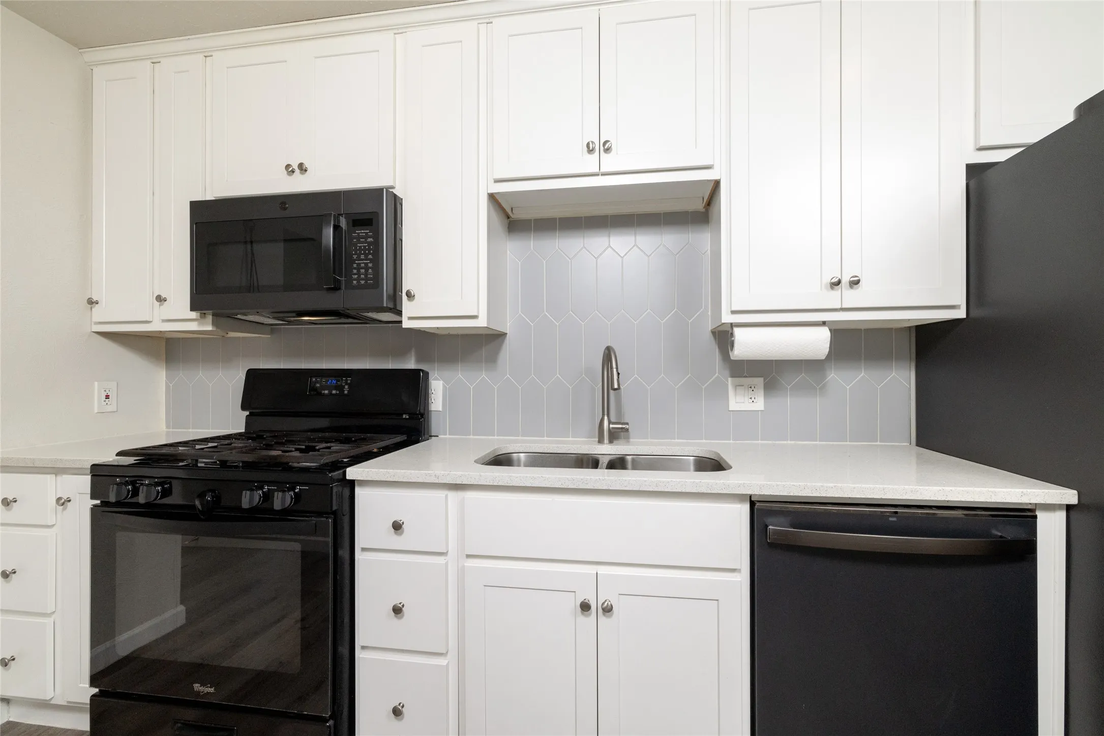White cabinetry, light-toned countertops, geometric tile backsplash, stainless steel sink, and black appliances