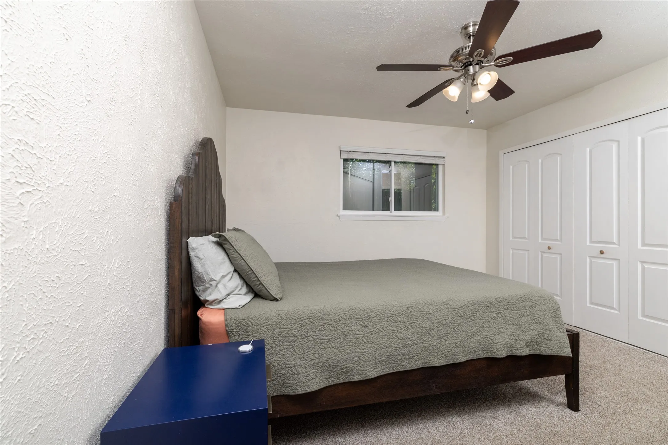 Textured wall finish, ceiling fan with integrated lighting, window with white trim, bi-fold closet doors, and neutral carpet flooring